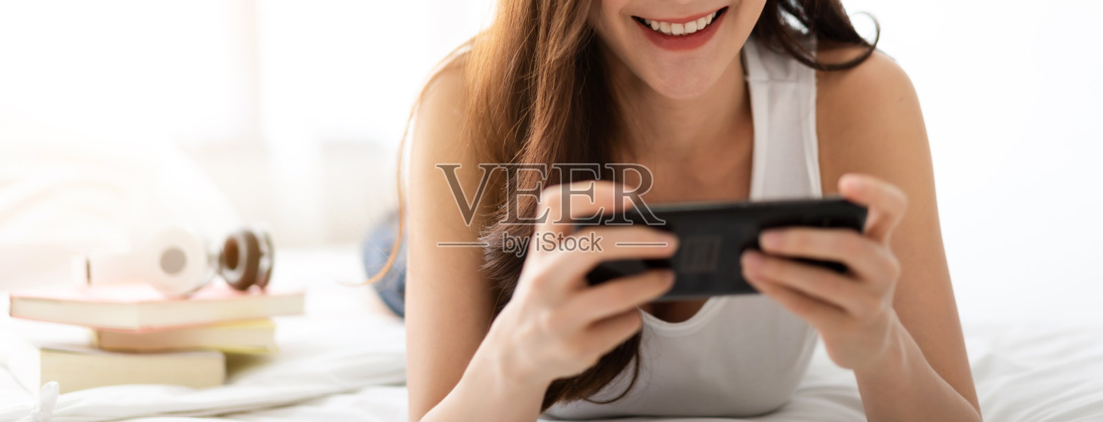 微笑的快乐开朗的美丽漂亮的亚洲女人放松地看着现代智能手机设备的技术。女孩喜欢在家里的床上玩手机和打字。科技照片摄影图片