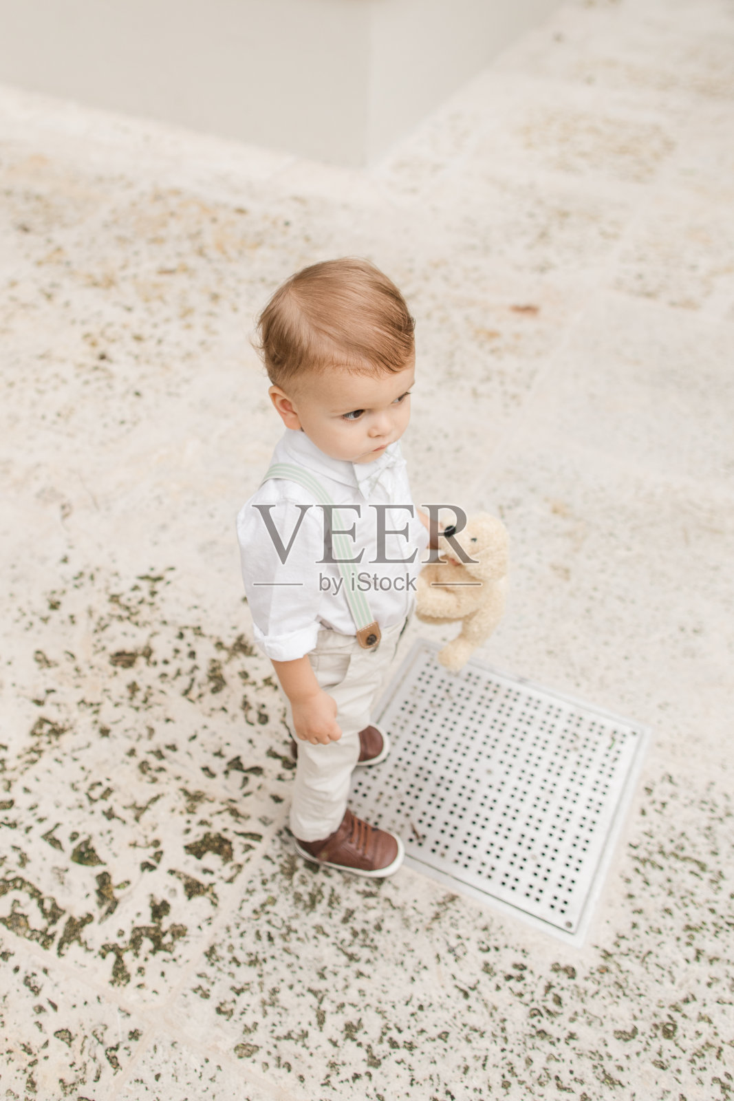 2023年5月，佛罗里达州热带棕榈滩，一个1岁的古巴裔美国小男孩，一头浅棕色短发，戴着鼠尾草绿色领结，穿着背带裤，白色纽扣衬衫，裤子和棕色鞋子，在花园里散步照片摄影图片