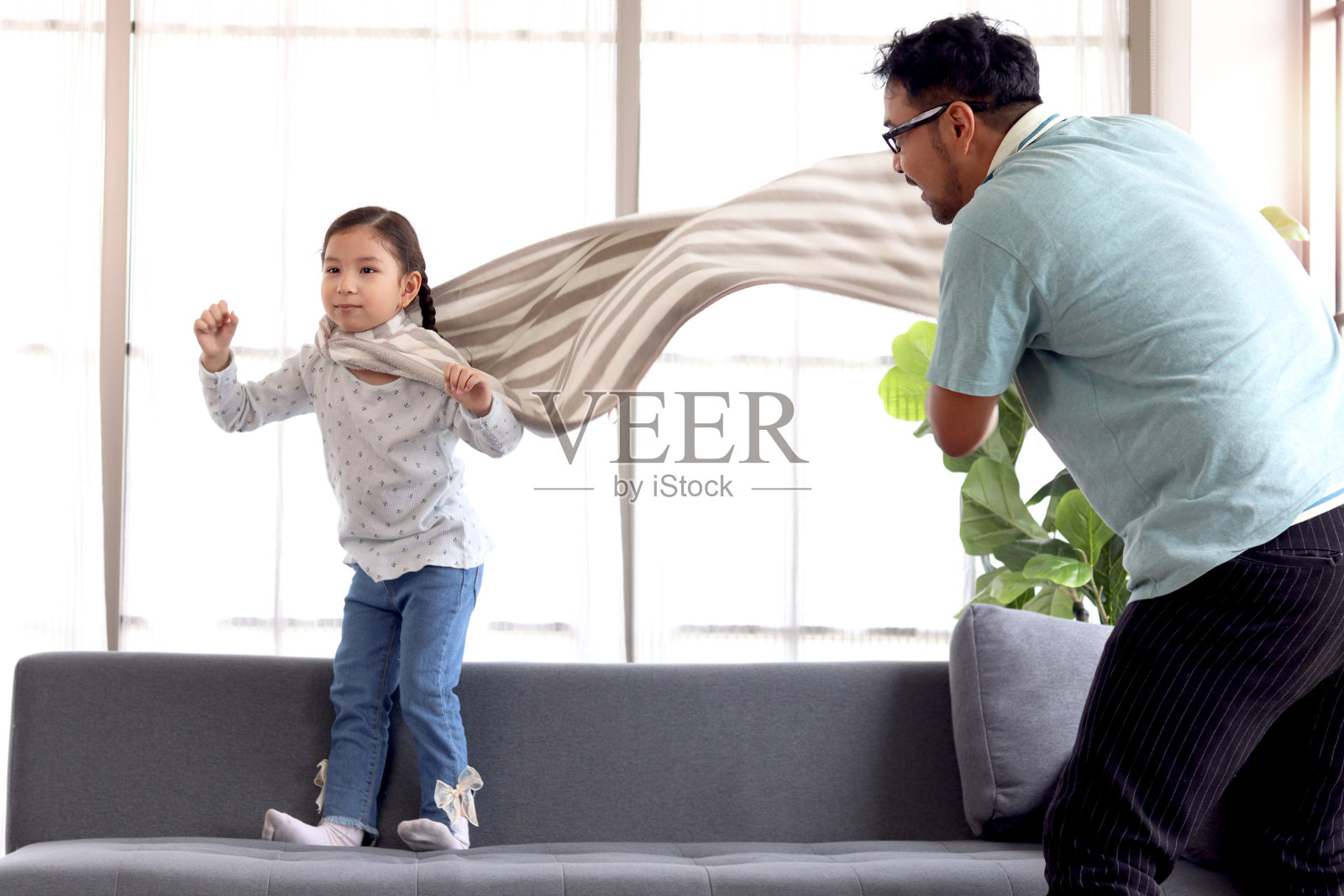 可爱的小女孩穿着大毛巾当超级英雄斗篷，爸爸抖着毛巾，假装她可以飞到客厅的沙发上，孩子和爸爸在家里一起扮演英雄，父亲节快乐。照片摄影图片