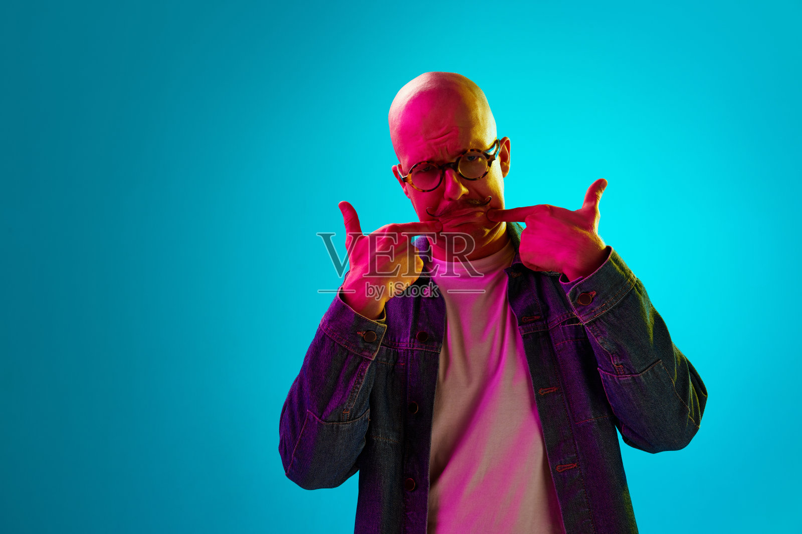 在霓虹灯的蓝色工作室背景下，留着小胡子的年轻秃顶男子的肖像。不愉快，不开心的脸照片摄影图片