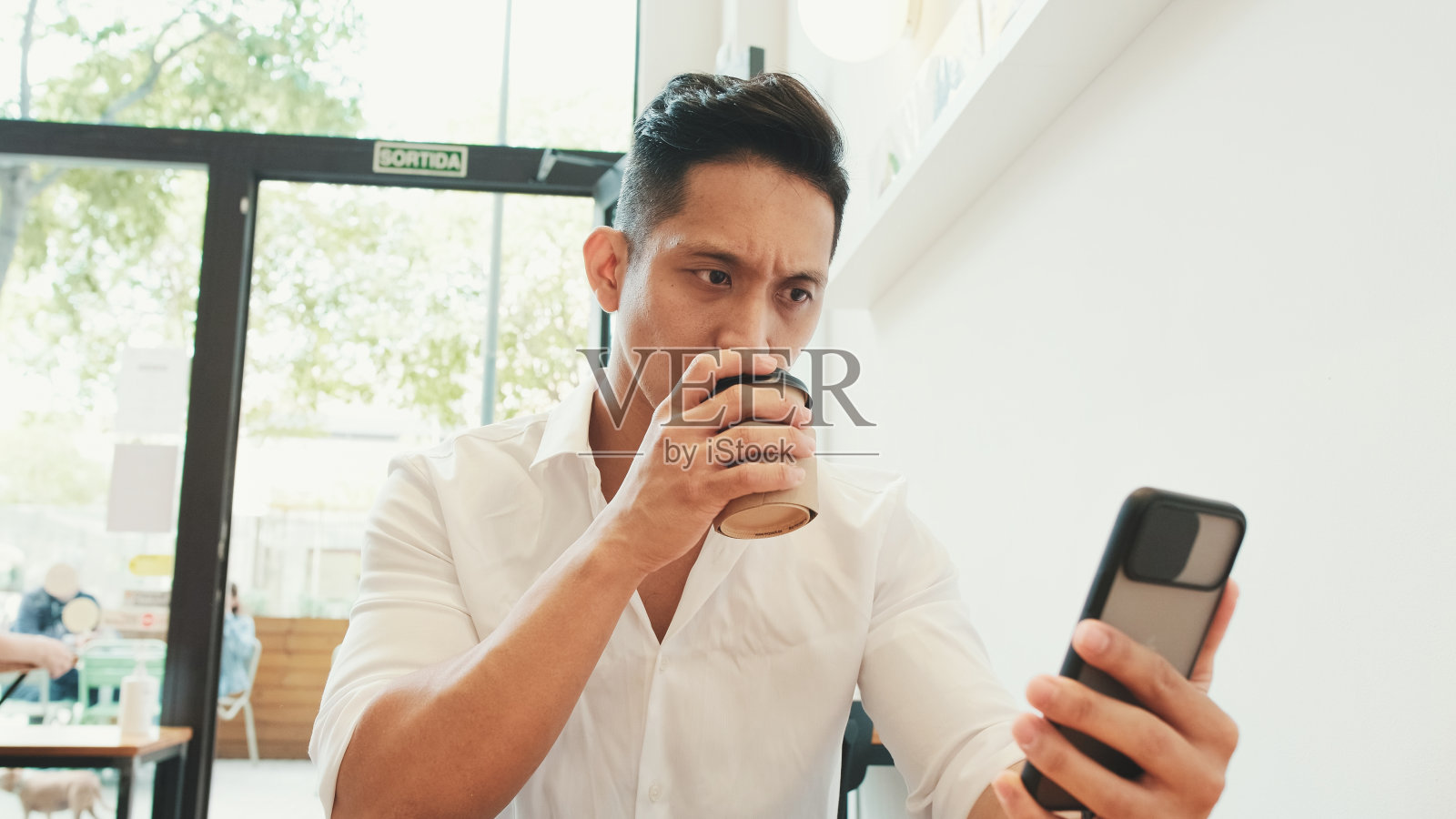 年轻的商人拿着手机坐在桌边，在咖啡馆里喝咖啡照片摄影图片