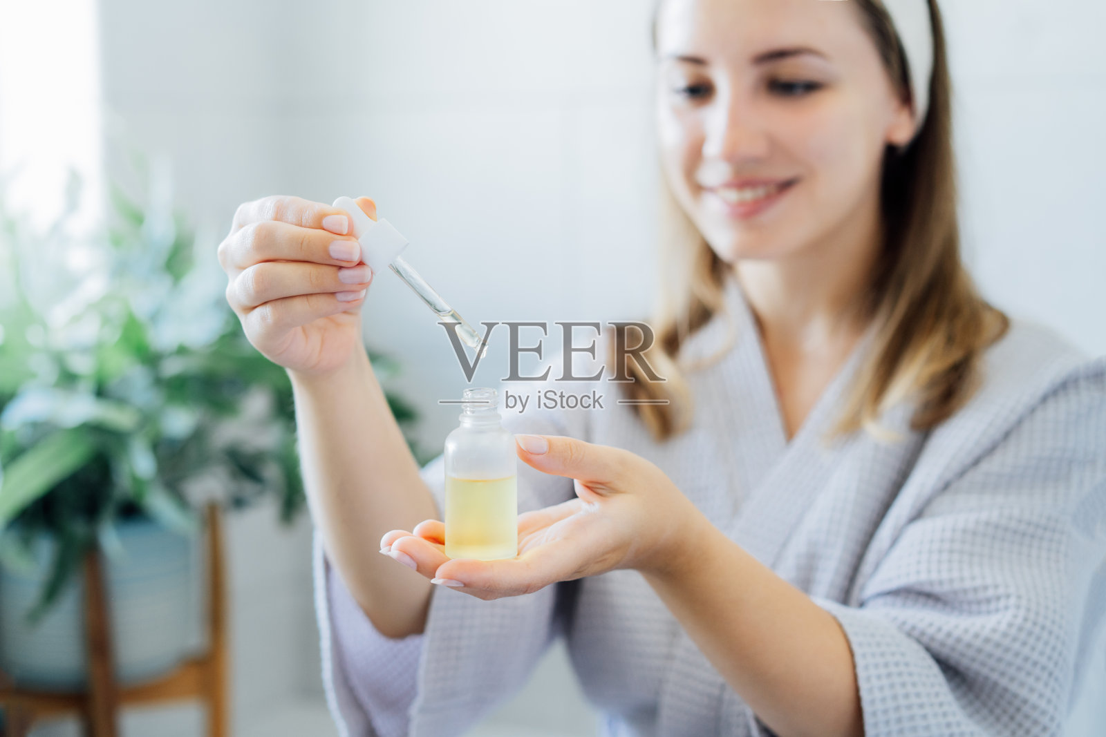 近距离观察穿着浴袍的年轻女子，手里拿着透明质酸血清的瓶子和滴管模型。皮肤保湿。美容水疗程序。居家美容自我护理。有选择性的重点照片摄影图片