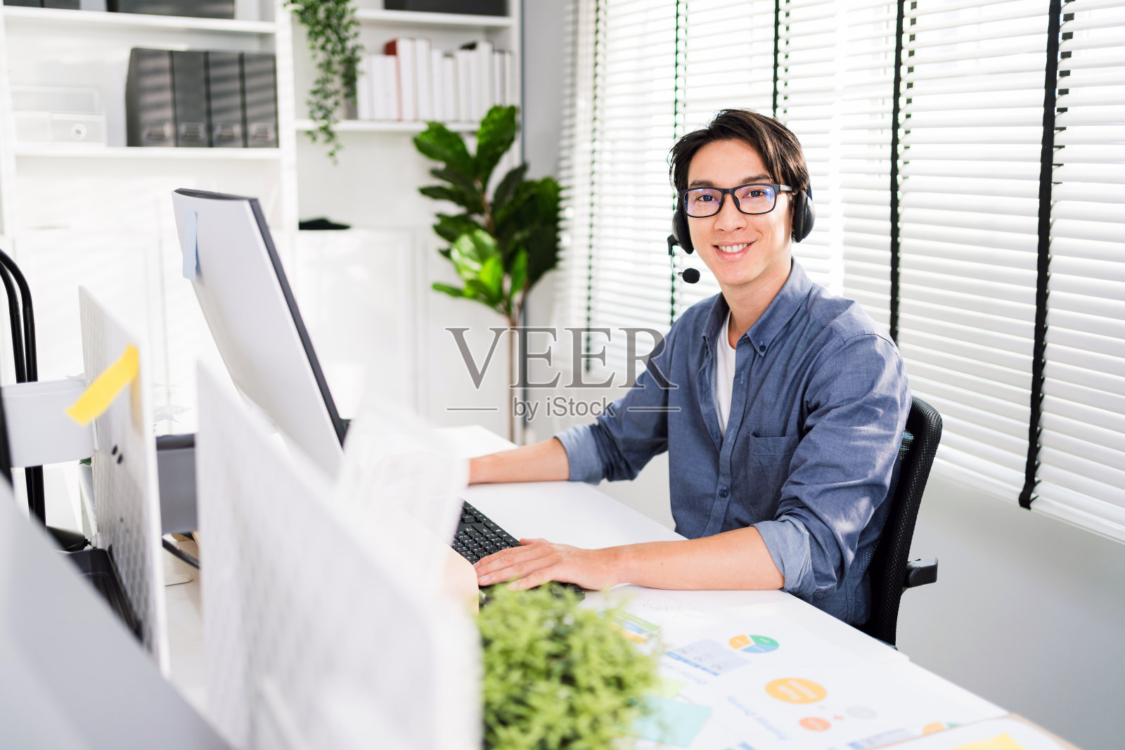 一位面带微笑的亚洲男子在呼叫中心担任客户支持接线员，戴着耳机。销售人员坐在办公桌前看着镜头的肖像。客户服务支持代表。照片摄影图片