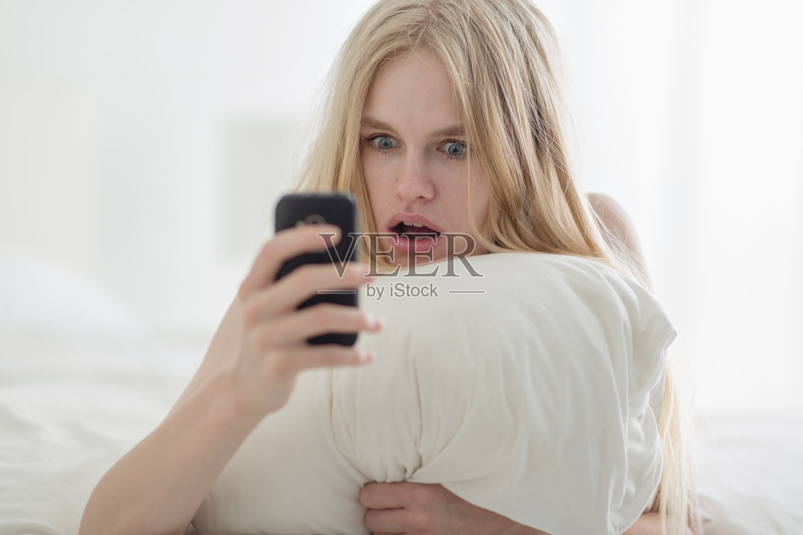 震惊的女人在床上看手机。照片摄影图片