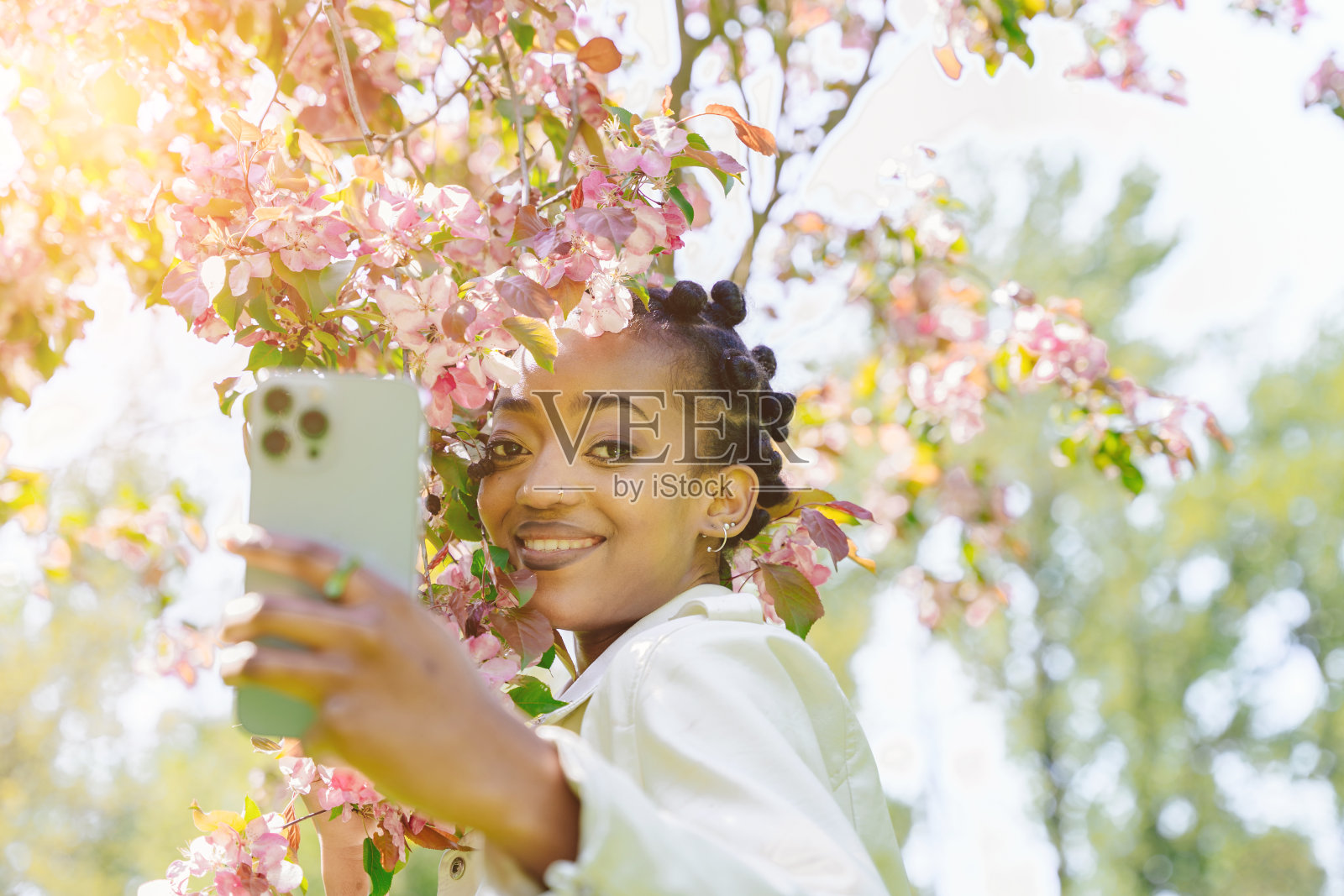 让我自拍一张。美丽的非洲女人看着手机，微笑着与盛开的粉红色鲜花树自拍的特写。照片摄影图片