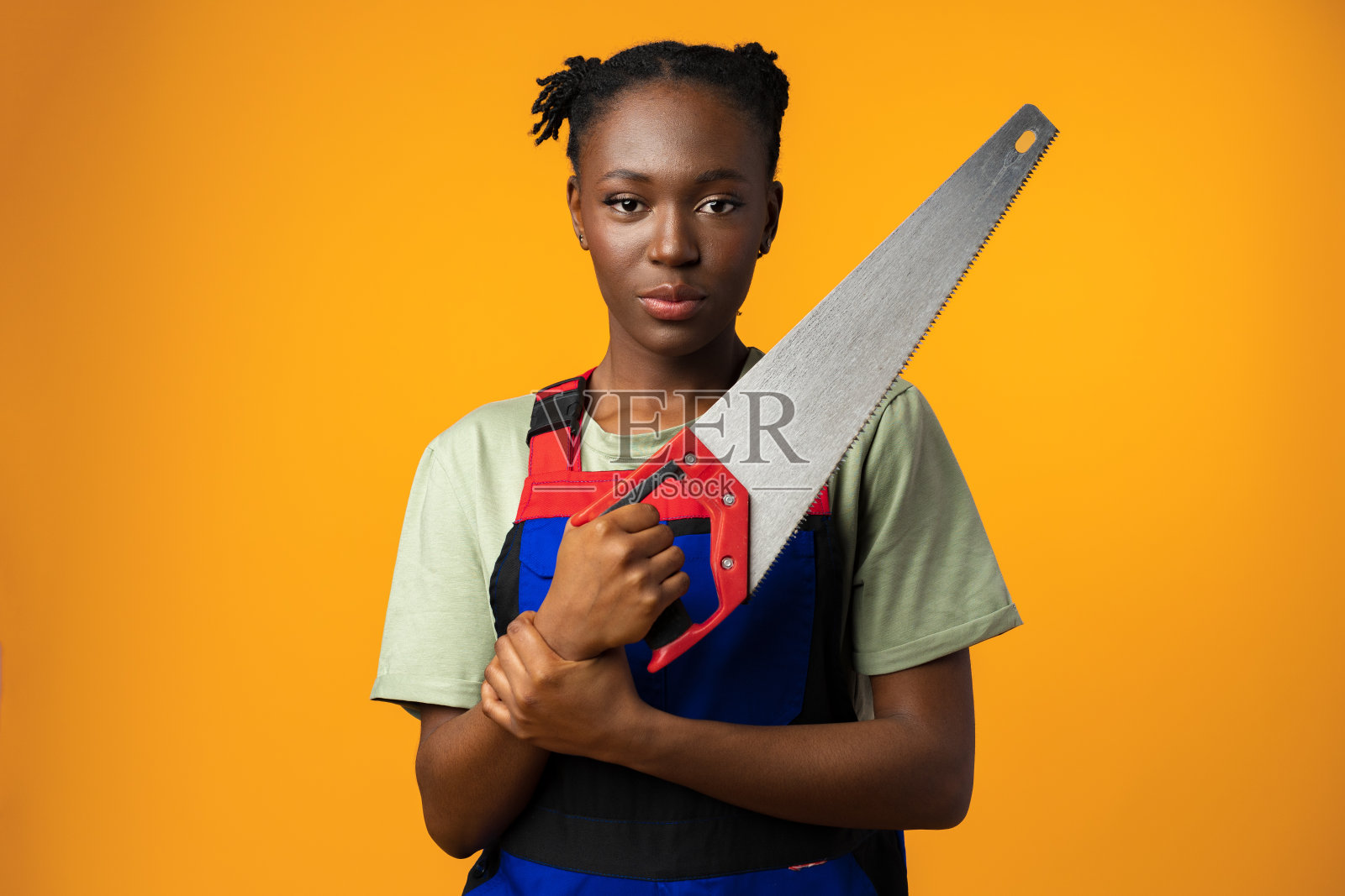 身穿制服的非洲裔美国妇女，在黄色的背景下，手里拿着锯子摆姿势照片摄影图片
