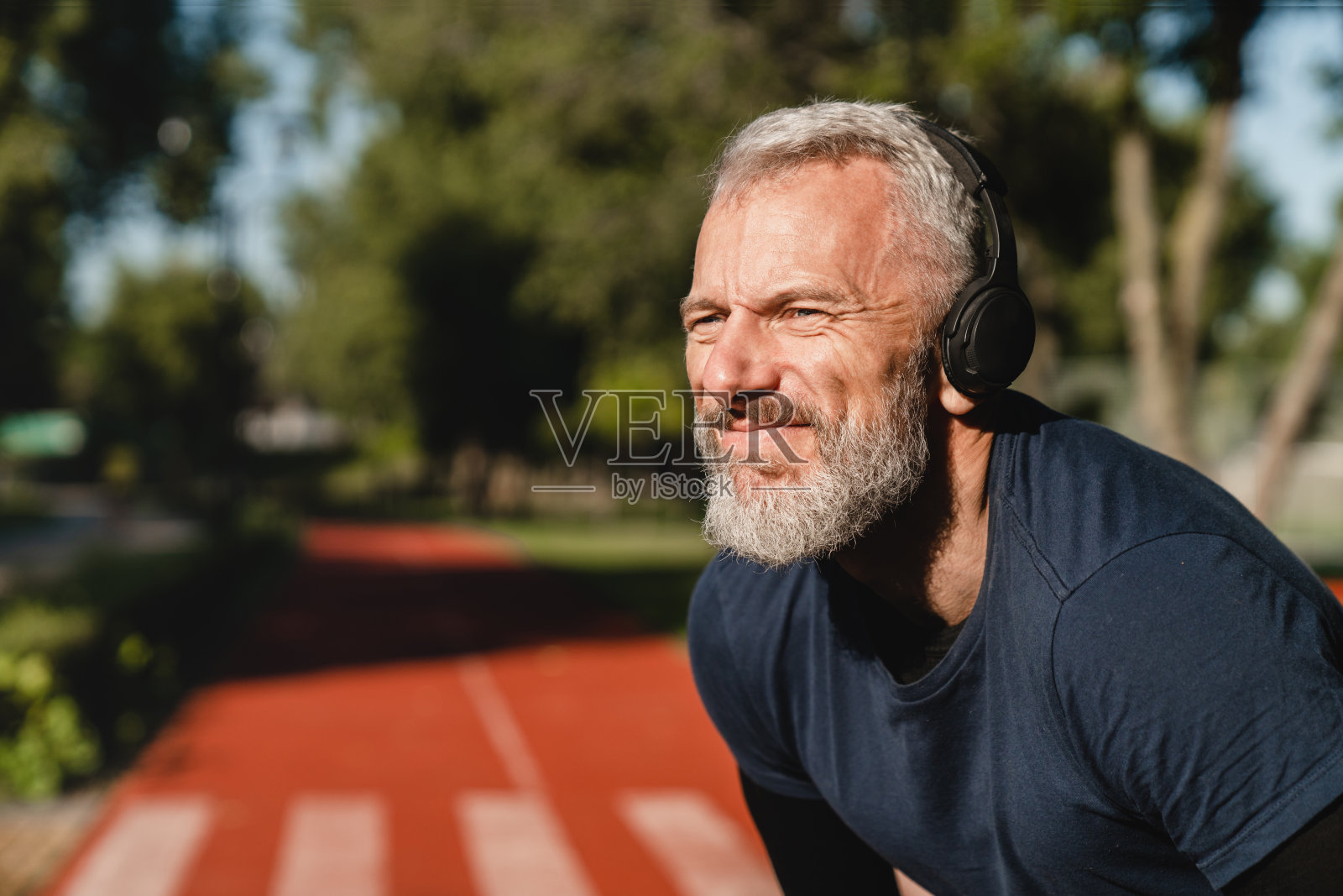 微笑英俊成熟的白人跑步者，戴着耳机听音乐，在体育场的户外训练。锻炼减肥。照片摄影图片