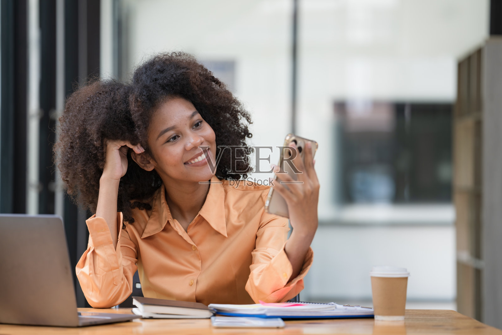 一个美丽自信的女商人坐在现代办公室里，拿着手机，拿着笔记本电脑。面带微笑的非裔美国自由职业者在家在线工作。照片摄影图片