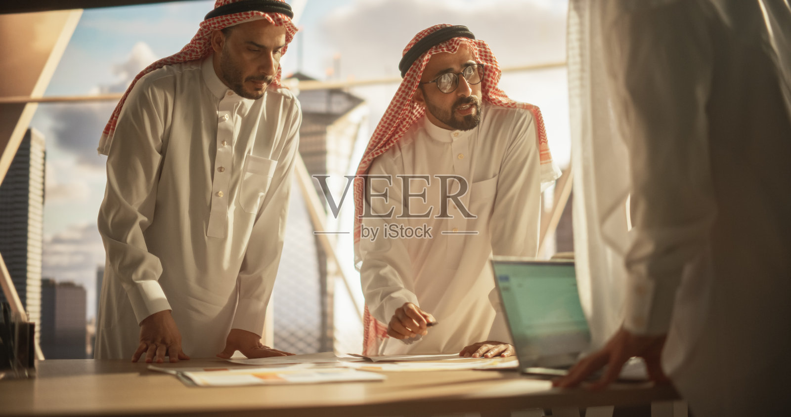 穿着白色传统服装的成功穆斯林商人正在办公室开会，谈判和谈论财务机会。使用笔记本电脑。沙特、阿联酋、阿拉伯商人概念。照片摄影图片