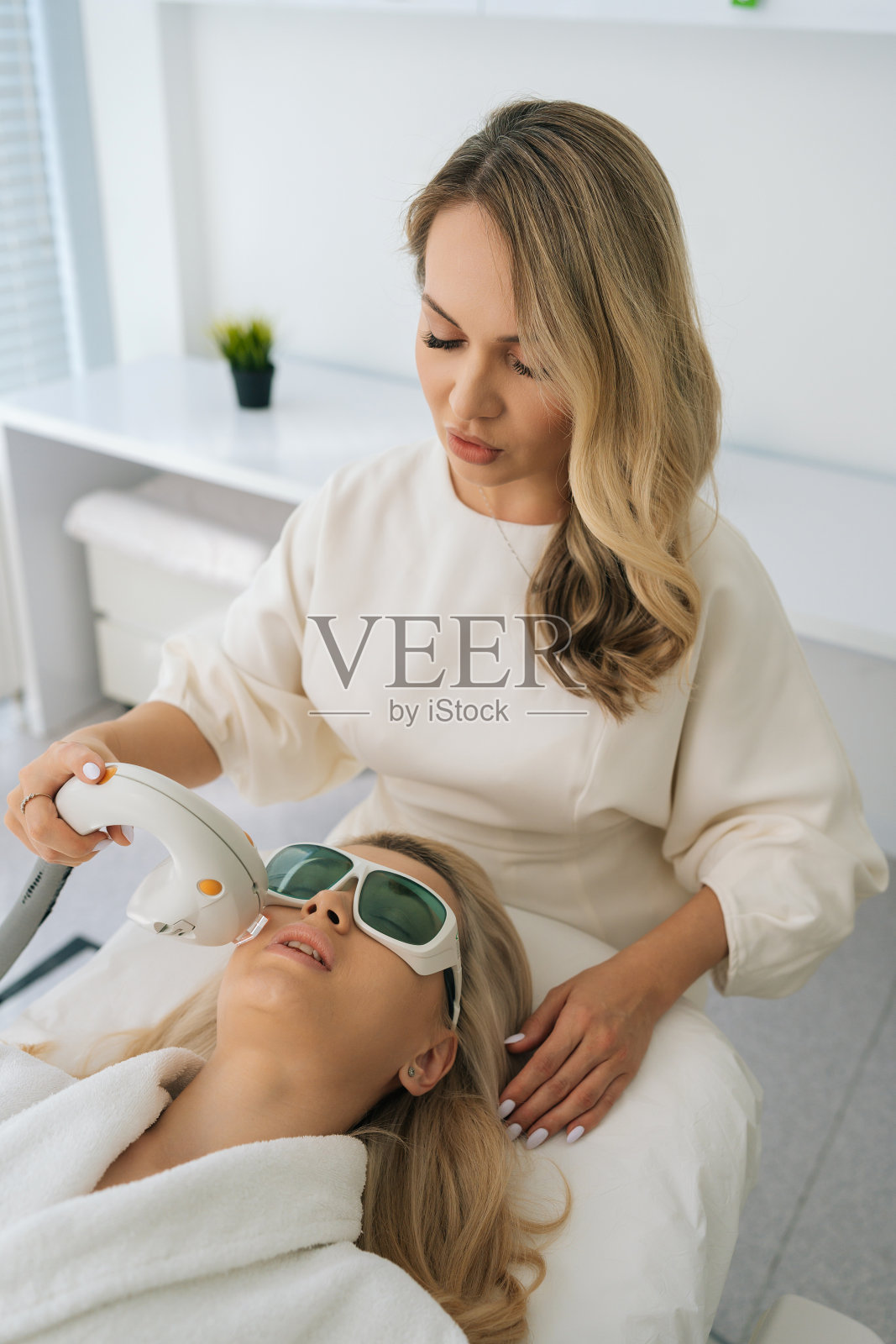 美容医学诊所女美容师为金发女客户进行抗衰老面部光疗的垂直高角度视角。照片摄影图片