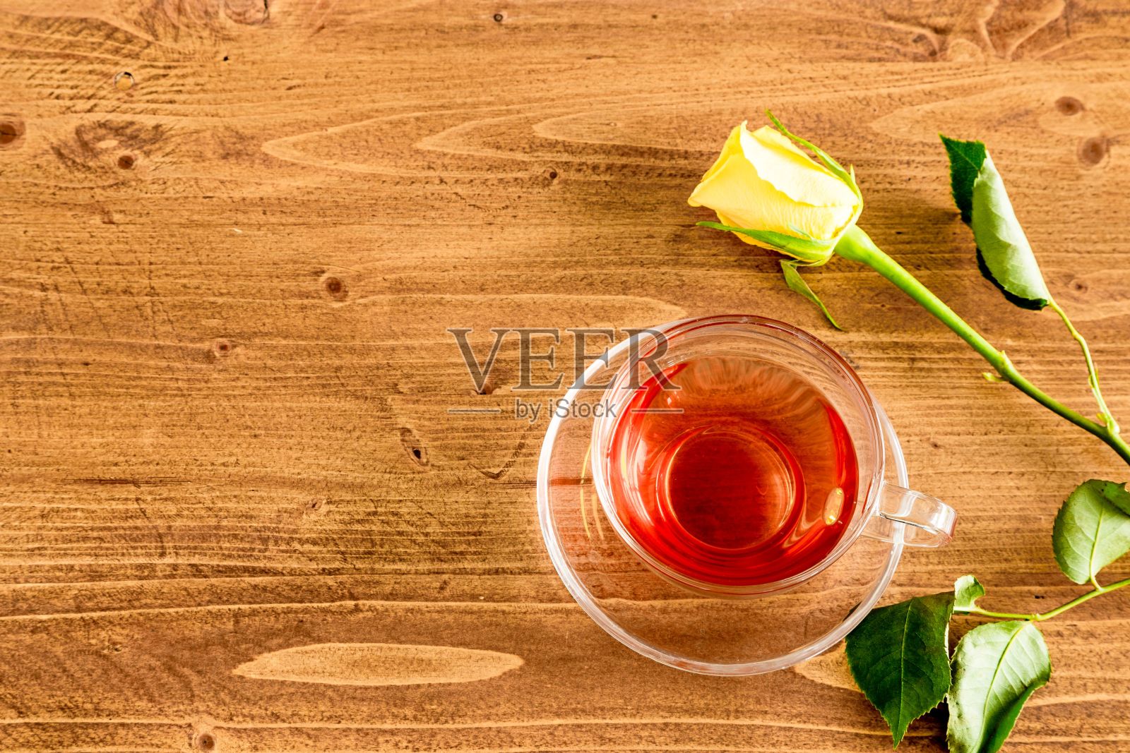 木桌上，一朵黄玫瑰和一杯装在透明玻璃杯里的红茶。照片摄影图片