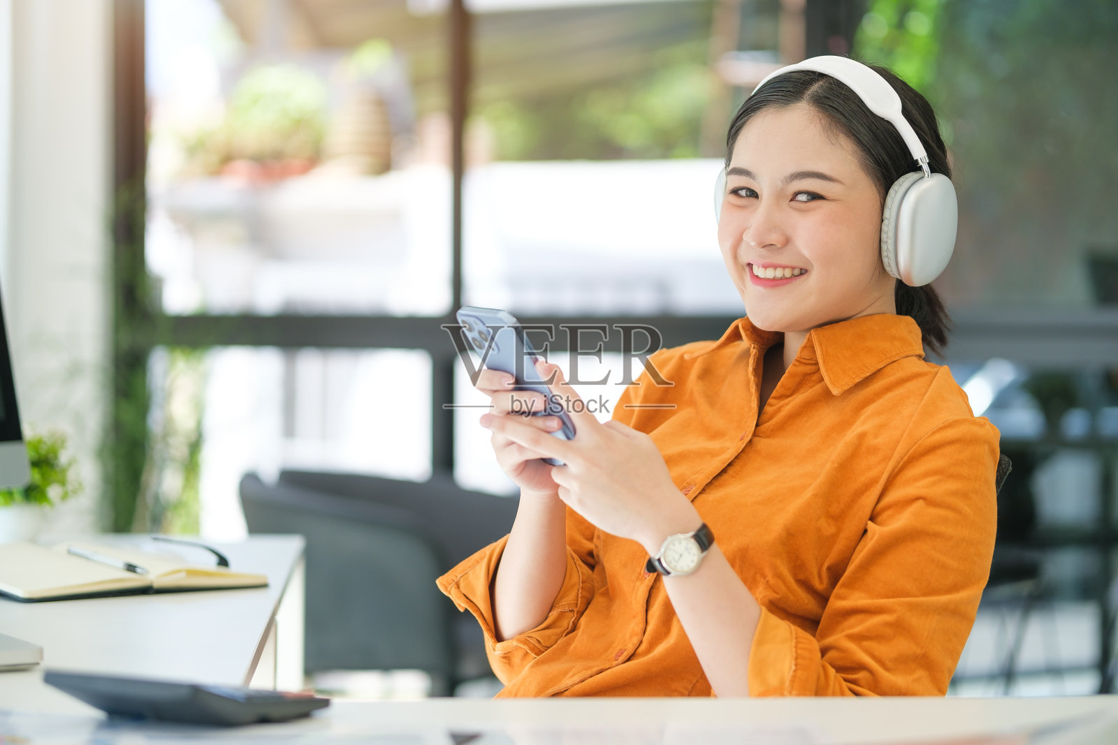 漂亮的年轻女子戴着无线耳机在明亮的现代办公室里使用手机。照片摄影图片