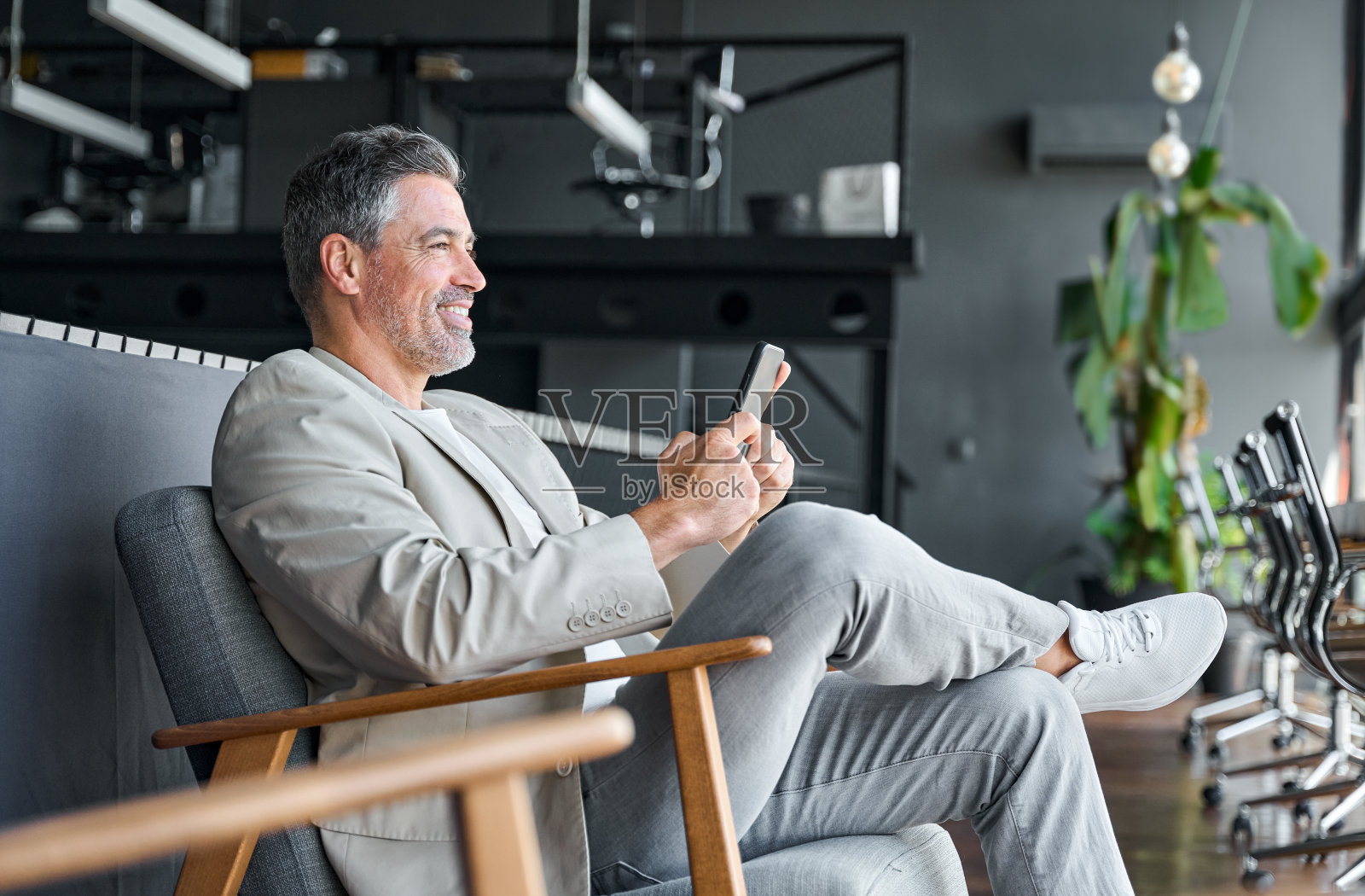 微笑的中年商人坐在办公室的椅子上使用手机。照片摄影图片