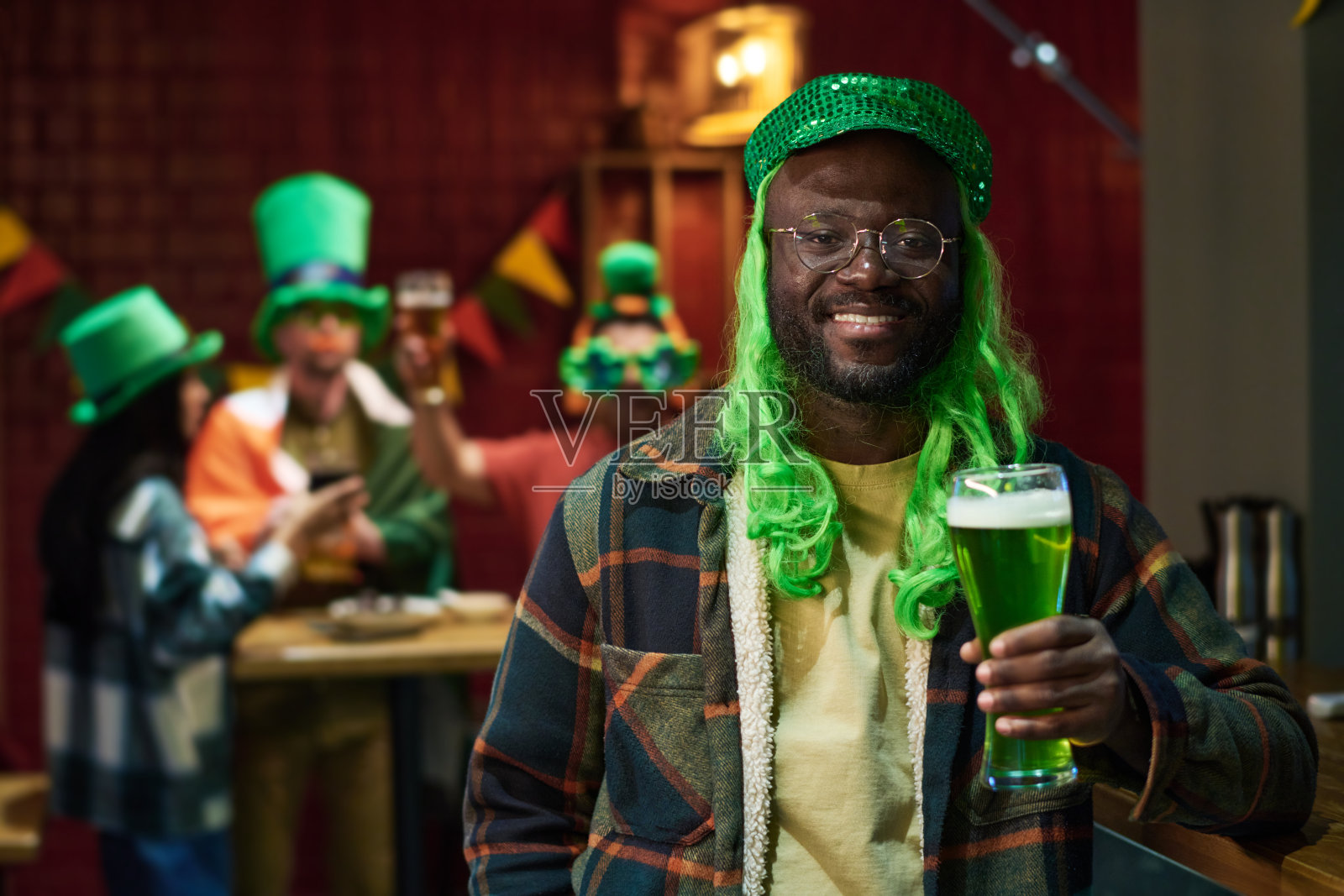 快乐的年轻非裔美国人戴着绿色的帽子和假发喝着啤酒照片摄影图片