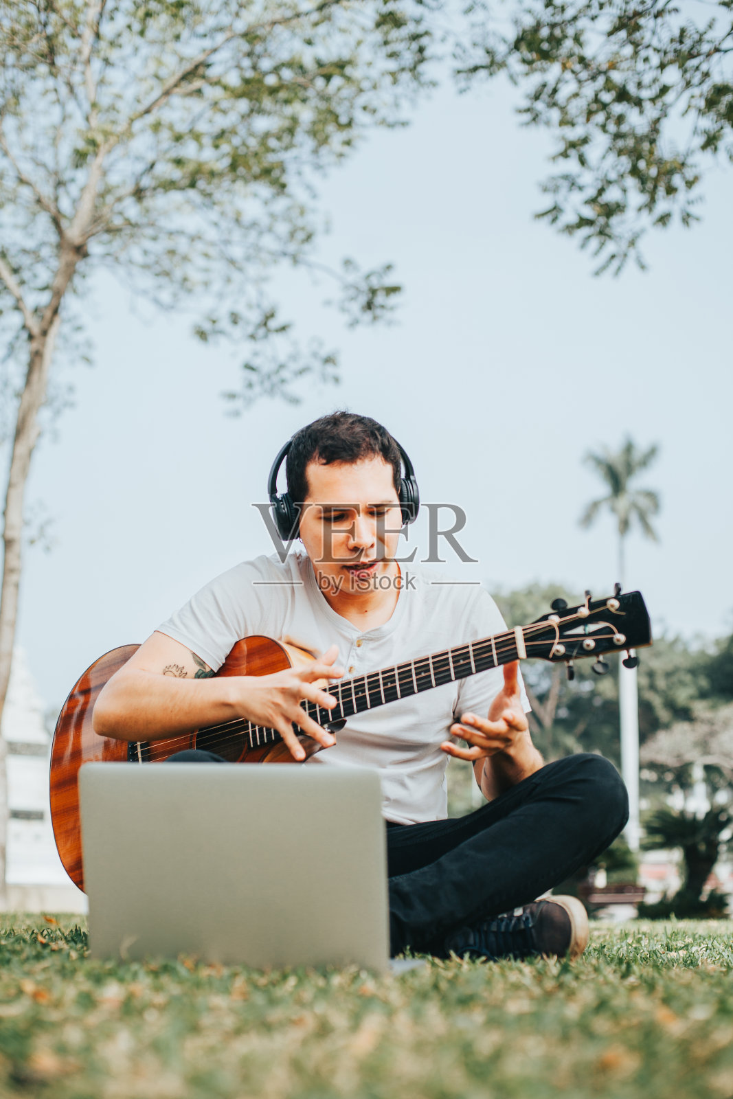 一个年轻人在公园里一边弹吉他一边用笔记本电脑教学。照片摄影图片