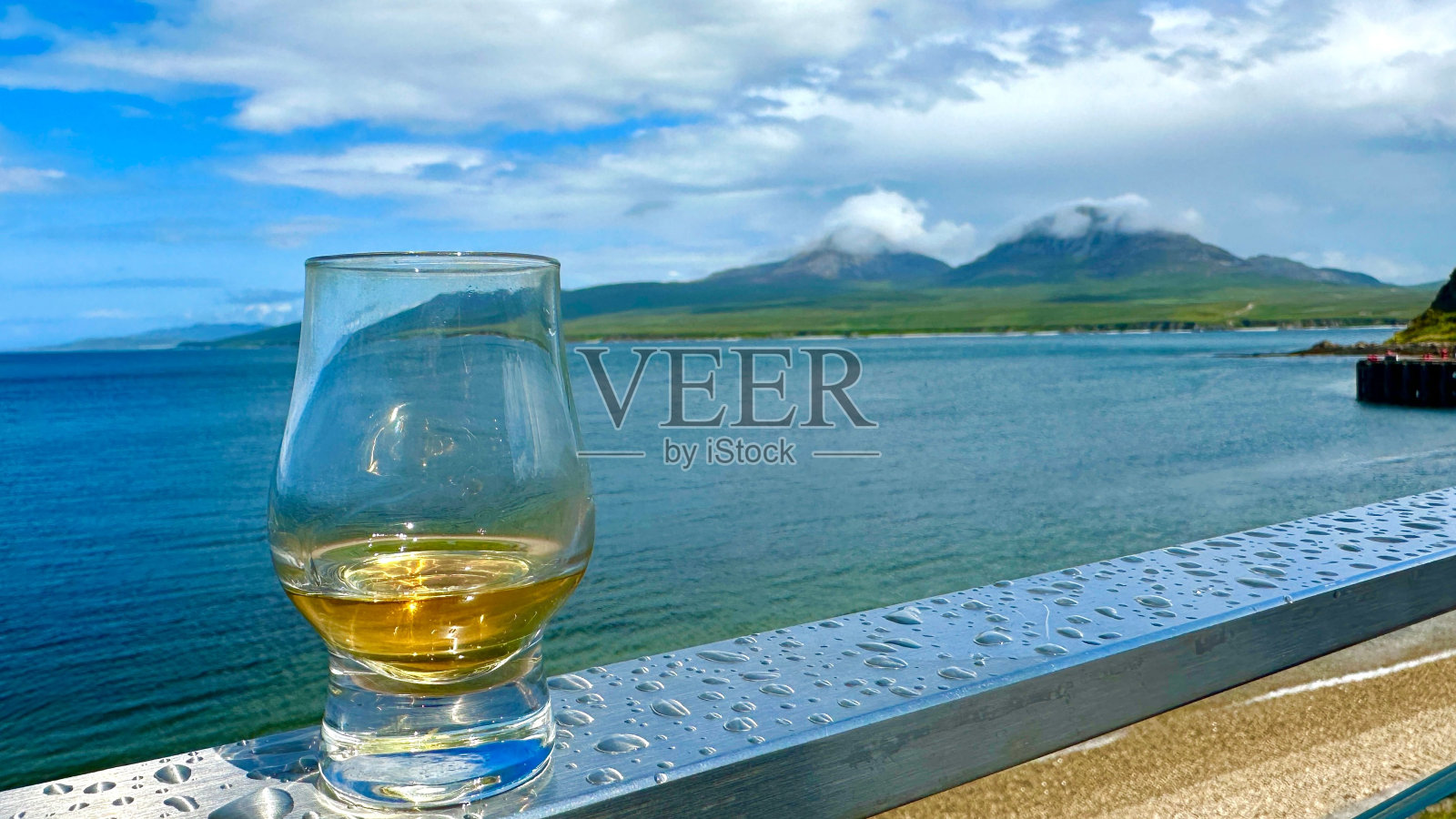 阳台栏杆上的一杯威士忌，背景是海洋和山脉的景色照片摄影图片