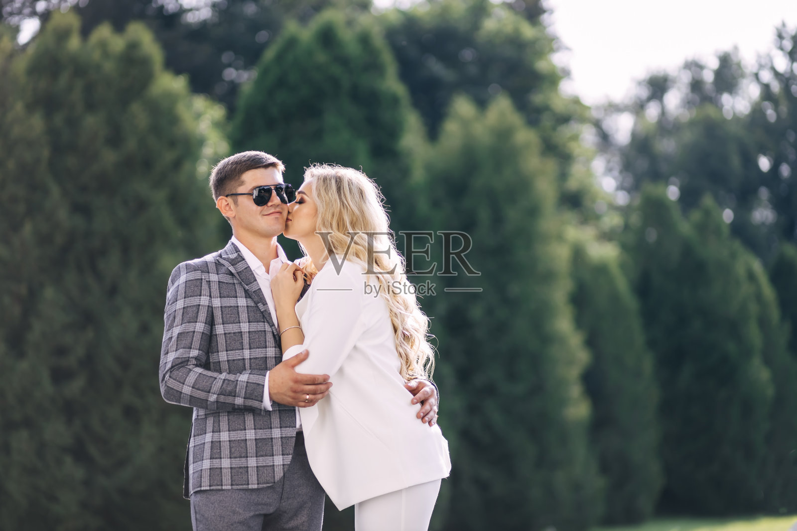 在以绿树为背景的公园里，一位身穿白色西装的美丽新娘亲吻着微笑的戴着眼镜的新郎的脸颊。热恋中的新婚夫妇。照片摄影图片