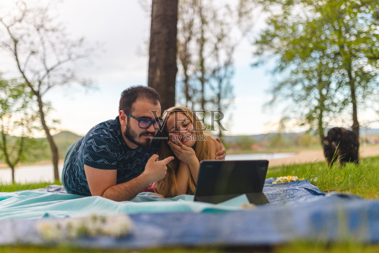 一对年轻夫妇躺在野外的野餐毯子上，同时用平板电脑进行视频通话照片摄影图片