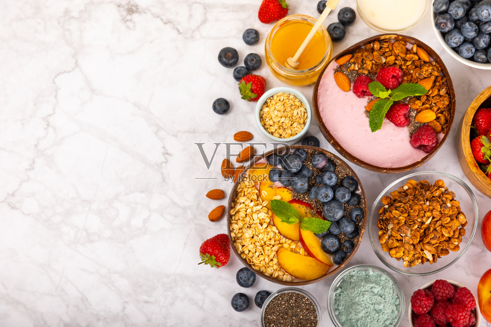 在厨房的桌子上，有一碗酸奶和新鲜的杏仁、蓝莓、覆盆子、桃子和草莓。巴西莓和螺旋藻碗。健康早餐概念。副本的空间。放文字的地方。照片摄影图片