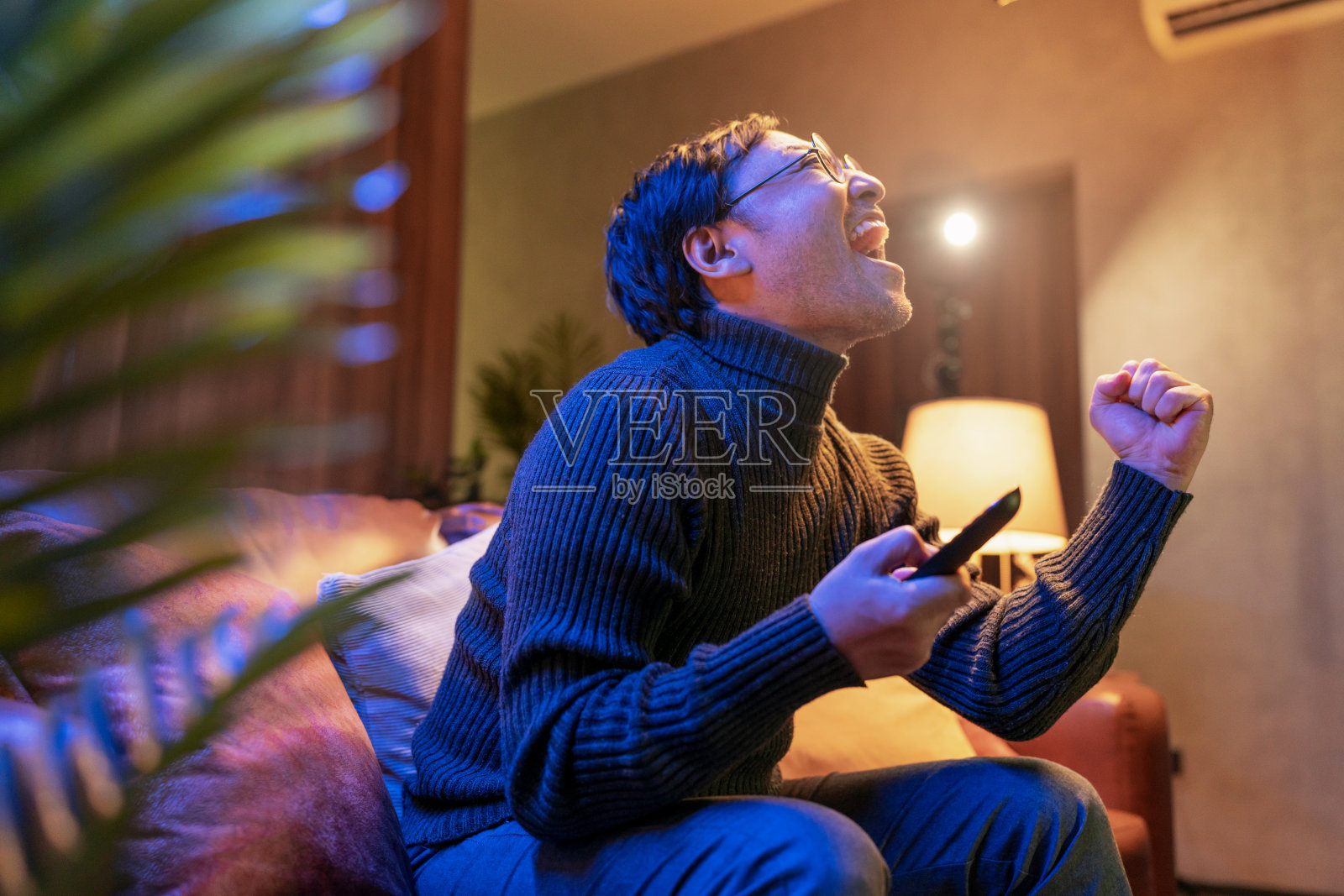一名亚洲成年男子坐在沙发上看电视上的体育比赛。一名兴奋的男子看着足球比赛，手举起来庆祝进球，在家里高声欢呼照片摄影图片