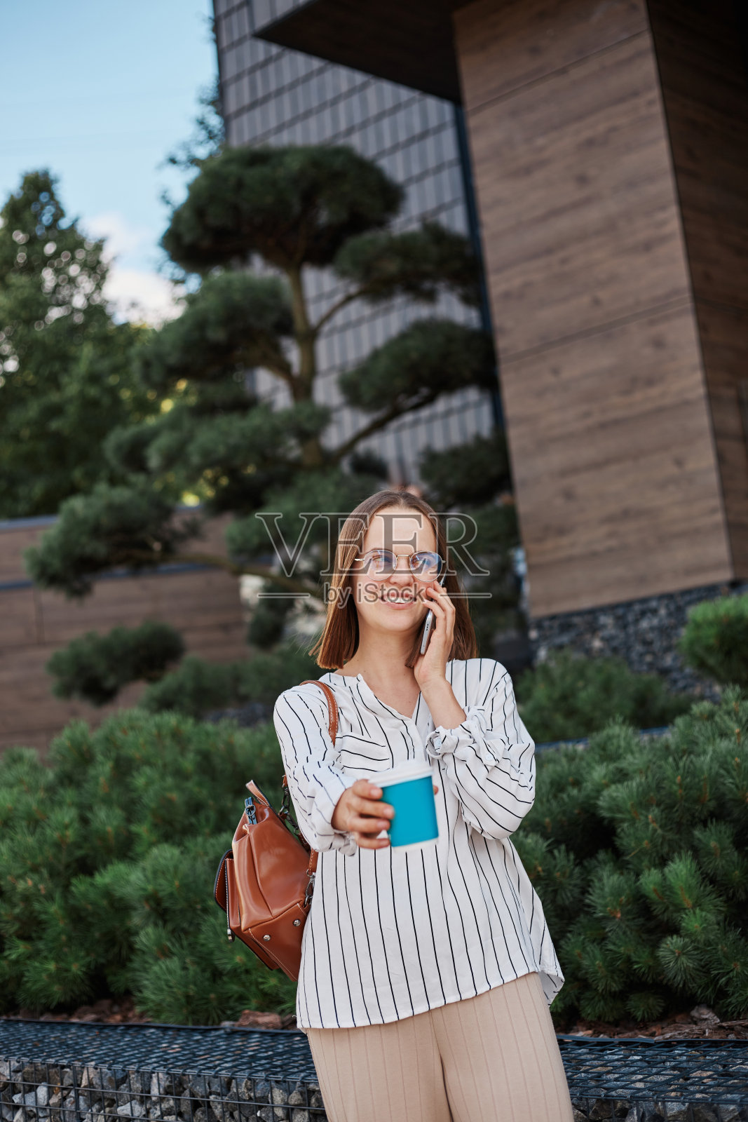 微笑迷人的积极乐观的女人穿着衬衫和裤子站在外卖咖啡杯和讲手机开心地笑。照片摄影图片