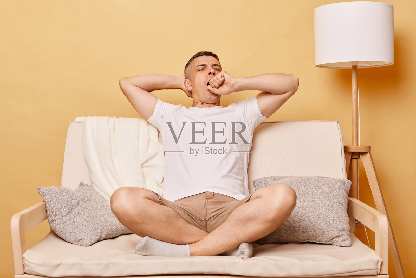 困倦疲惫的黑发成年男子穿着休闲服，坐在沙发上靠着米色的墙，感觉疲惫打着哈欠，用手掌捂住嘴。照片摄影图片