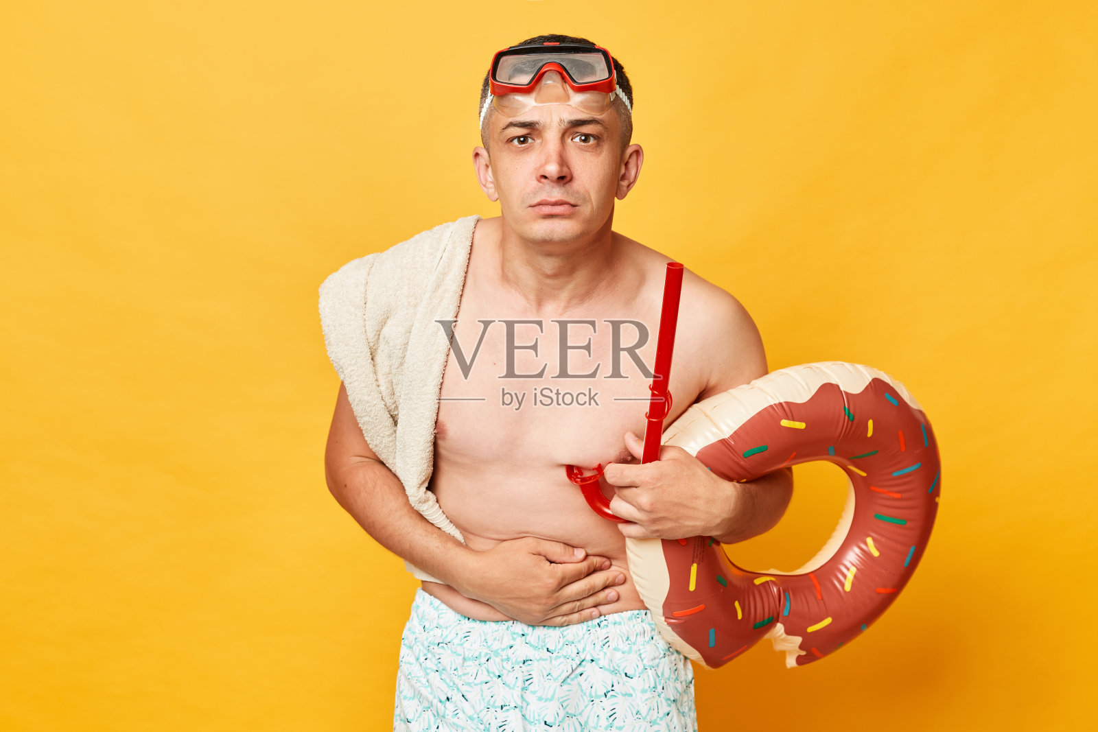 生病不健康的成年男子穿着短裤，泳衣，潜水镜，拿着甜甜圈，橡胶圈和袋子孤立在黄色的背景感到肚子痛，皱眉的脸。照片摄影图片