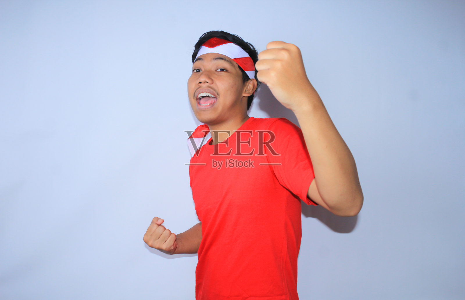 图为身穿红色t恤，头戴头巾的印尼青年在独立日高举拳头庆祝成功或进球。年轻人对自己的成就和满足做出“是”的手势照片摄影图片