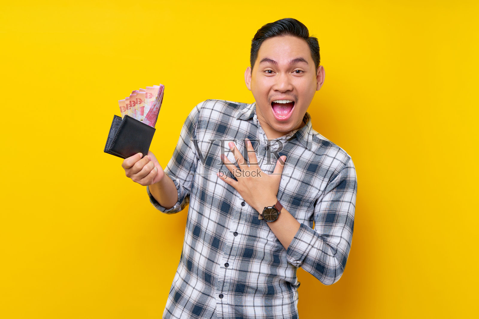 惊讶的年轻英俊的亚洲男子穿着白色格子衬衫，手里拿着一个黄色背景的钱包，里面有现金。人的生活理念照片摄影图片