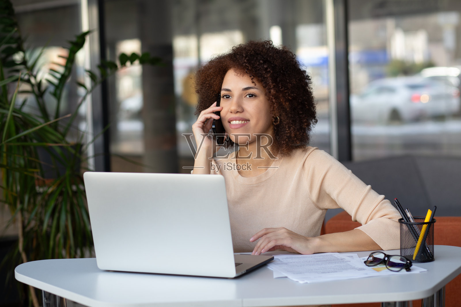 一个美丽自信的女商人的肖像，坐在现代办公室里，用笔记本电脑和手机聊天。照片摄影图片
