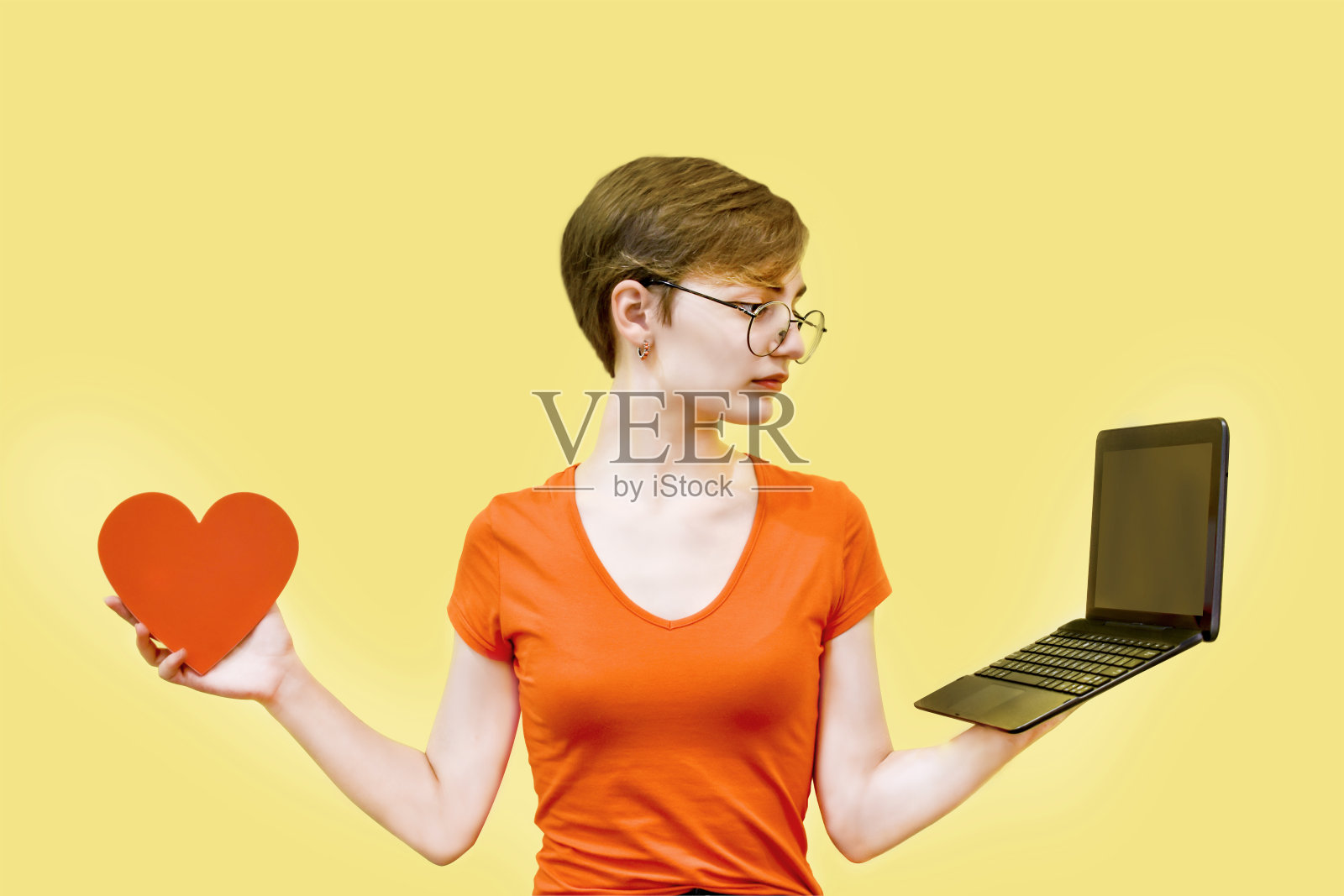 黄色背景的年轻女子一只手拿着笔记本，另一只手拿着心脏。选择的概念照片摄影图片