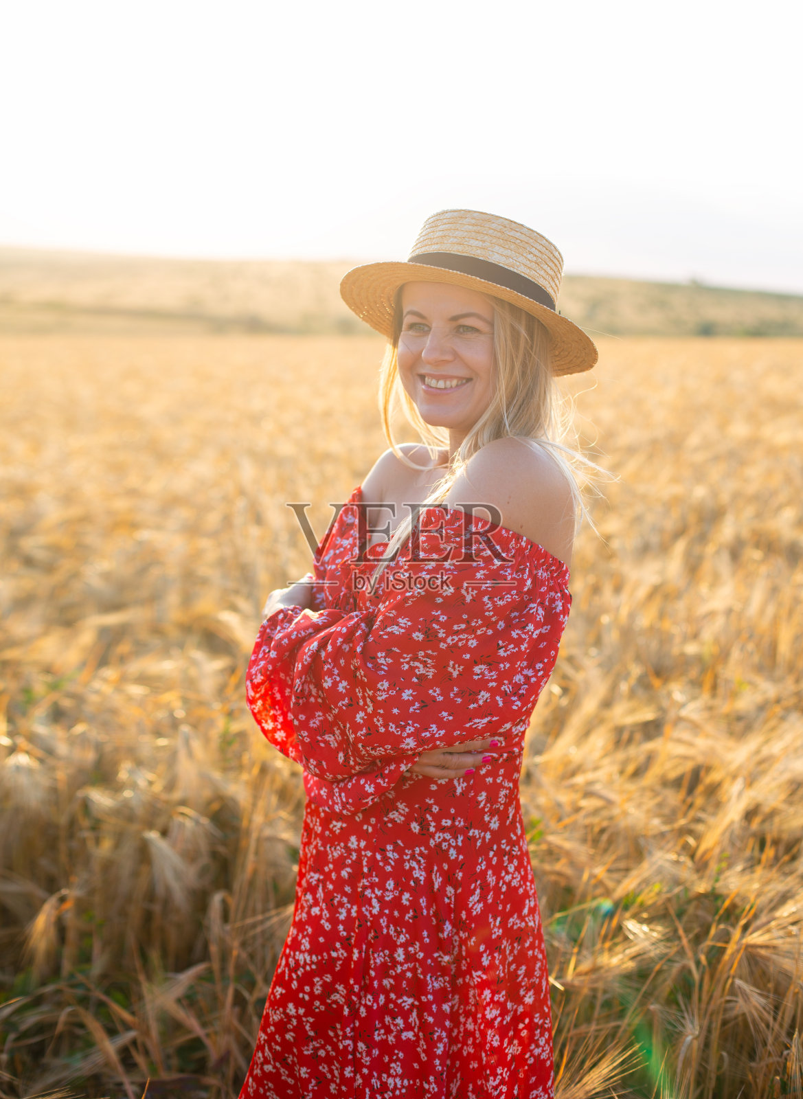 白种金发女人，穿着红裙子，戴着红帽子，站在夕阳下金色的麦田里照片摄影图片