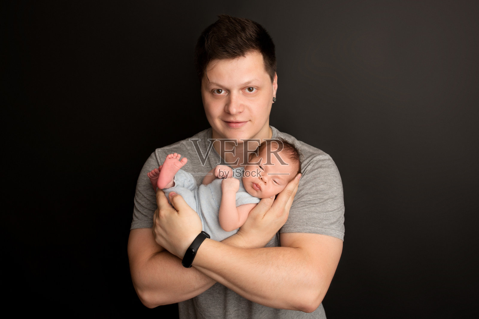 一个可爱的新生男孩在生命的最初几天穿着蓝色工装裤睡觉。在父亲的怀抱里。温暖的父母拥抱。黑色背景。专业人像摄影。照片摄影图片