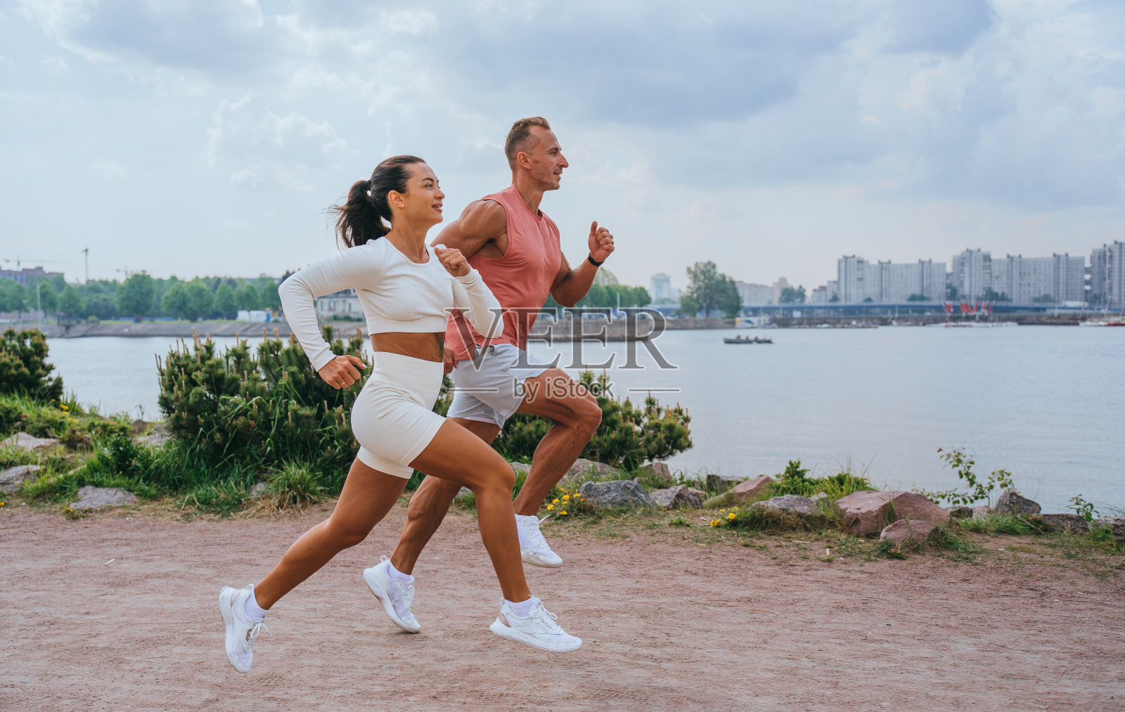 黝黑的美国年轻夫妇在公园河堤边跑步，可以看到河流和城市。深色皮肤的白人女人和丈夫在外面慢跑。男性健身教练与客户一起锻炼。照片摄影图片