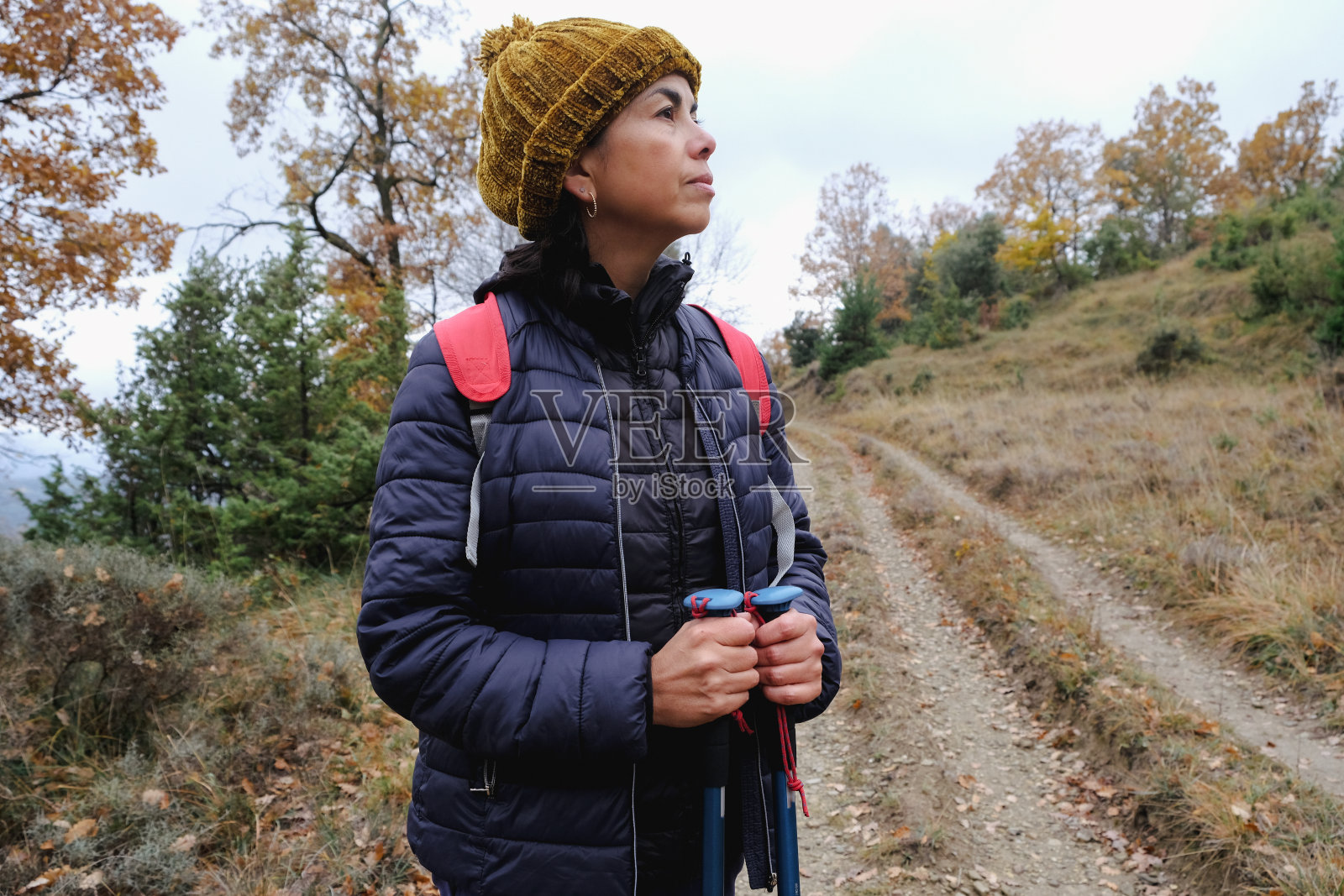 秋天，在西班牙的一个村庄里，一名妇女拿着登山杖，戴着冬季针织帽，在山间徒步旅行，享受着寂静和宁静照片摄影图片