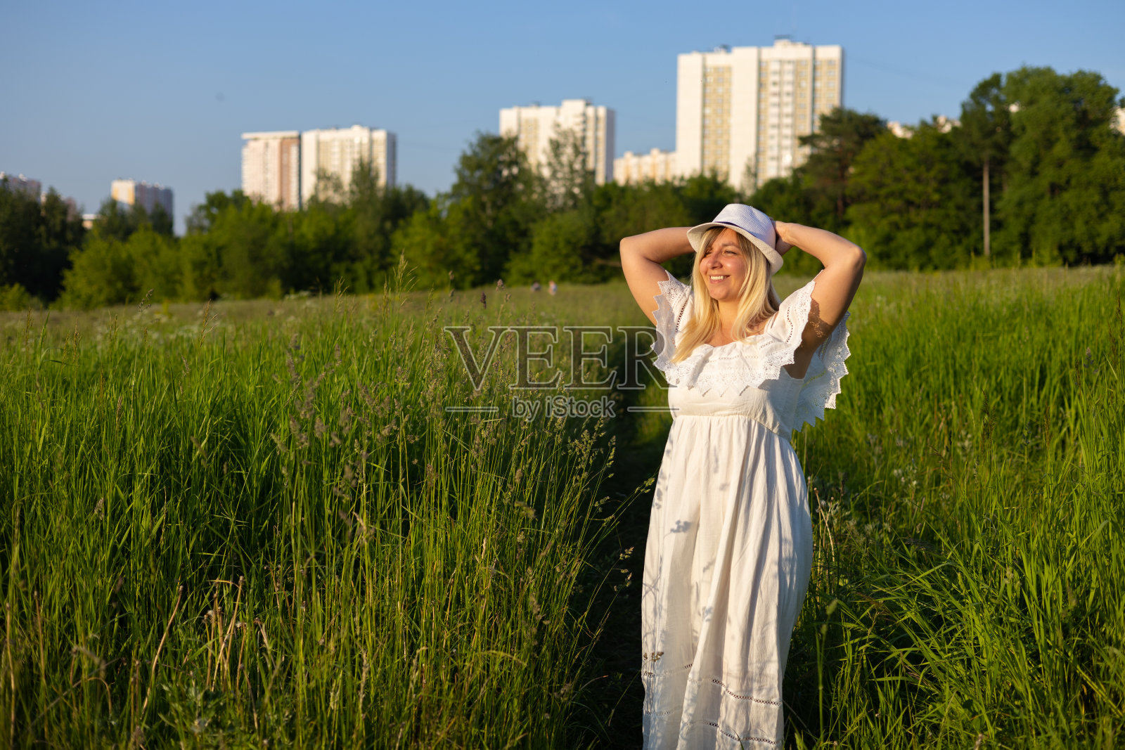 夏天，一名妇女在城市高楼大厦的背景下，在田野上享受夕阳的余晖。一位身穿白色背心裙的美女沐浴在落日的余晖中。照片摄影图片