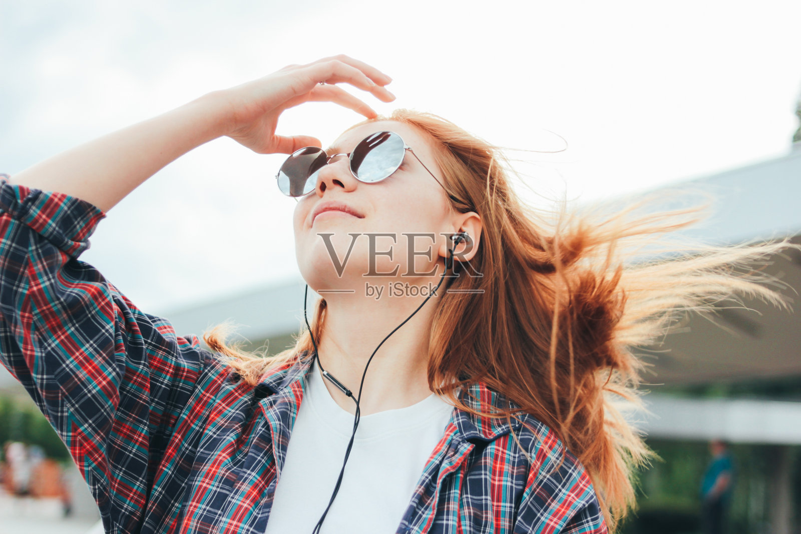 迷人的红发微笑的女孩，戴着圆形太阳镜，手里拿着手机，穿着休闲服，在城市的街道上听音乐照片摄影图片