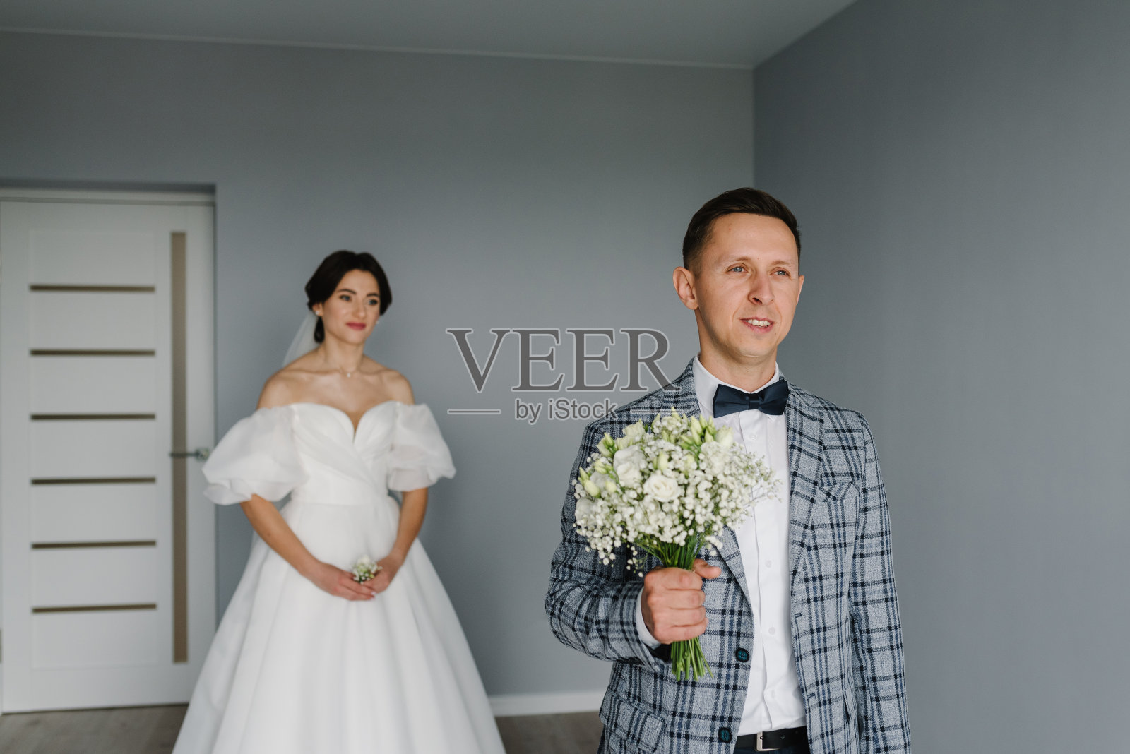 第一次见面是新婚夫妇。新娘回到新郎身边，惊喜在家。英俊的新郎在白色的房间里捧着一束花。新郎在等新娘。前视图。新婚快乐。照片摄影图片