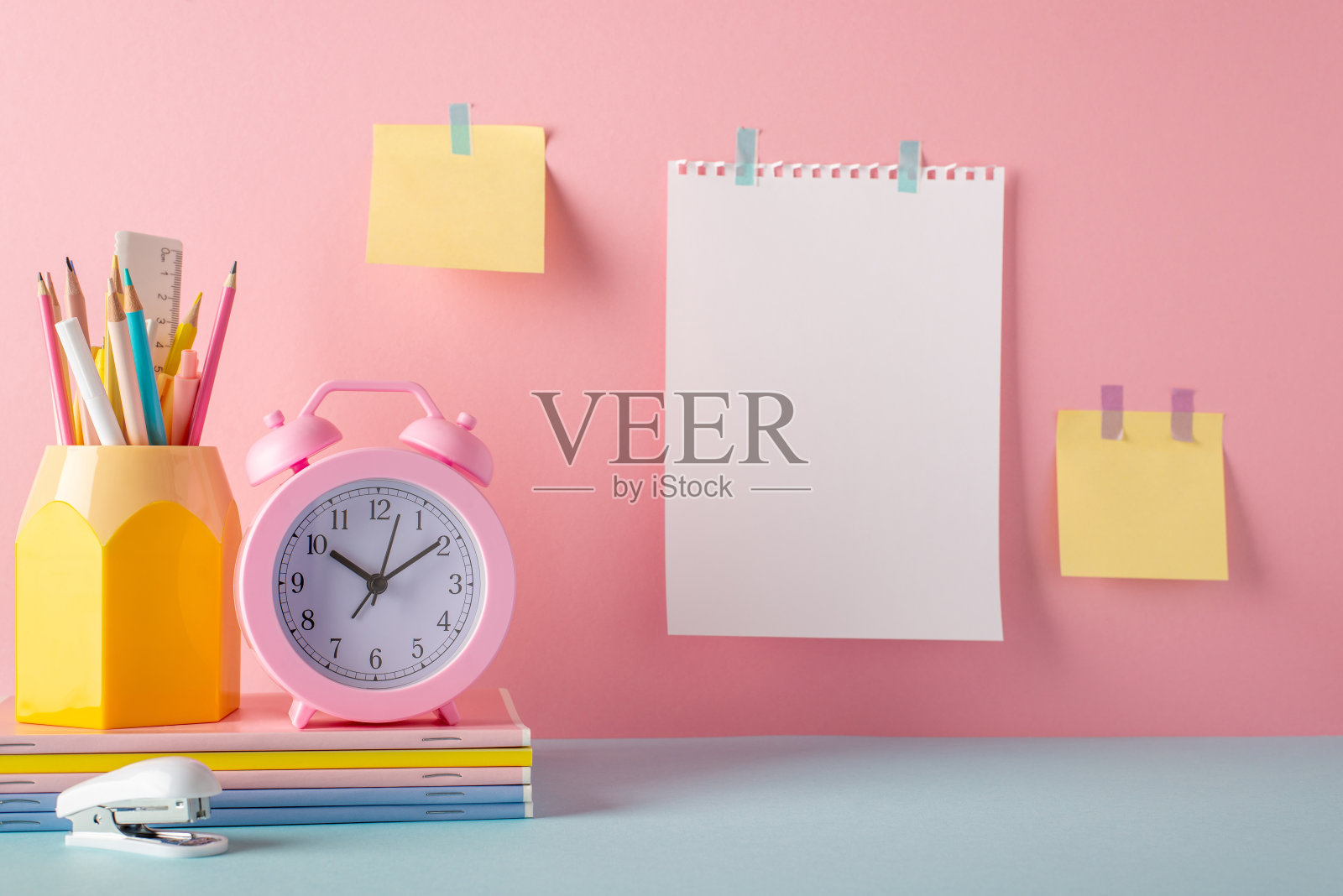学校用品概念。笔筒、闹钟、笔记本、订书机、便签和纸的照片，贴在粉红色的墙上，留白照片摄影图片