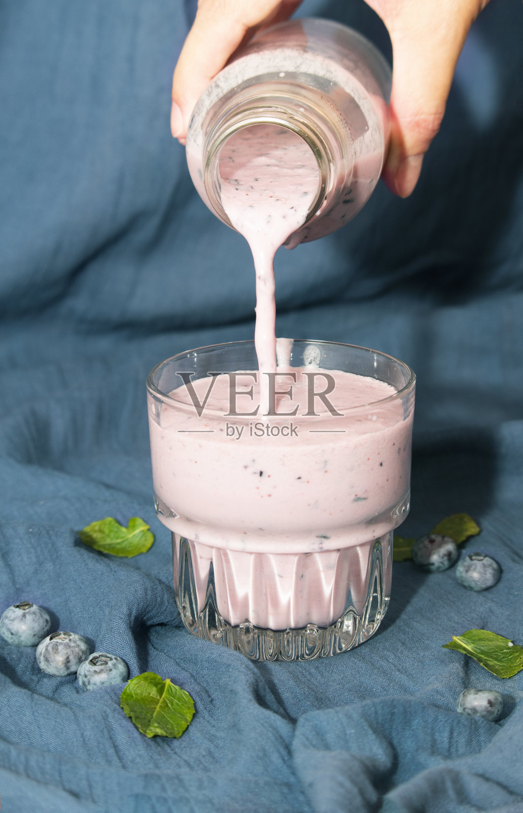 一位女士的手将一瓶蓝莓冰沙从瓶子里倒进玻璃杯里。排毒饮料照片摄影图片