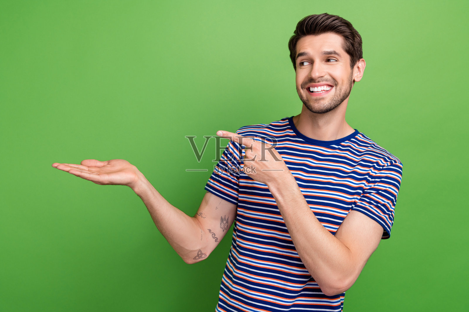 照片中迷人开朗的男子穿着条纹t恤，手指指着手臂，空旷的空间孤立绿色背景照片摄影图片