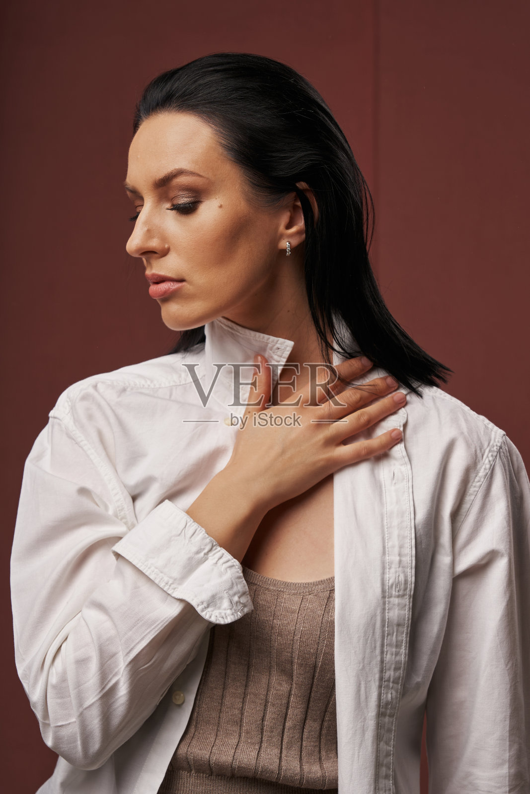 一个美丽的年轻女子的肖像，黑色光滑的头发，穿着白衬衫，谁转过身去，闭上眼睛，抓住衬衫领子在一个朴素的背景照片摄影图片