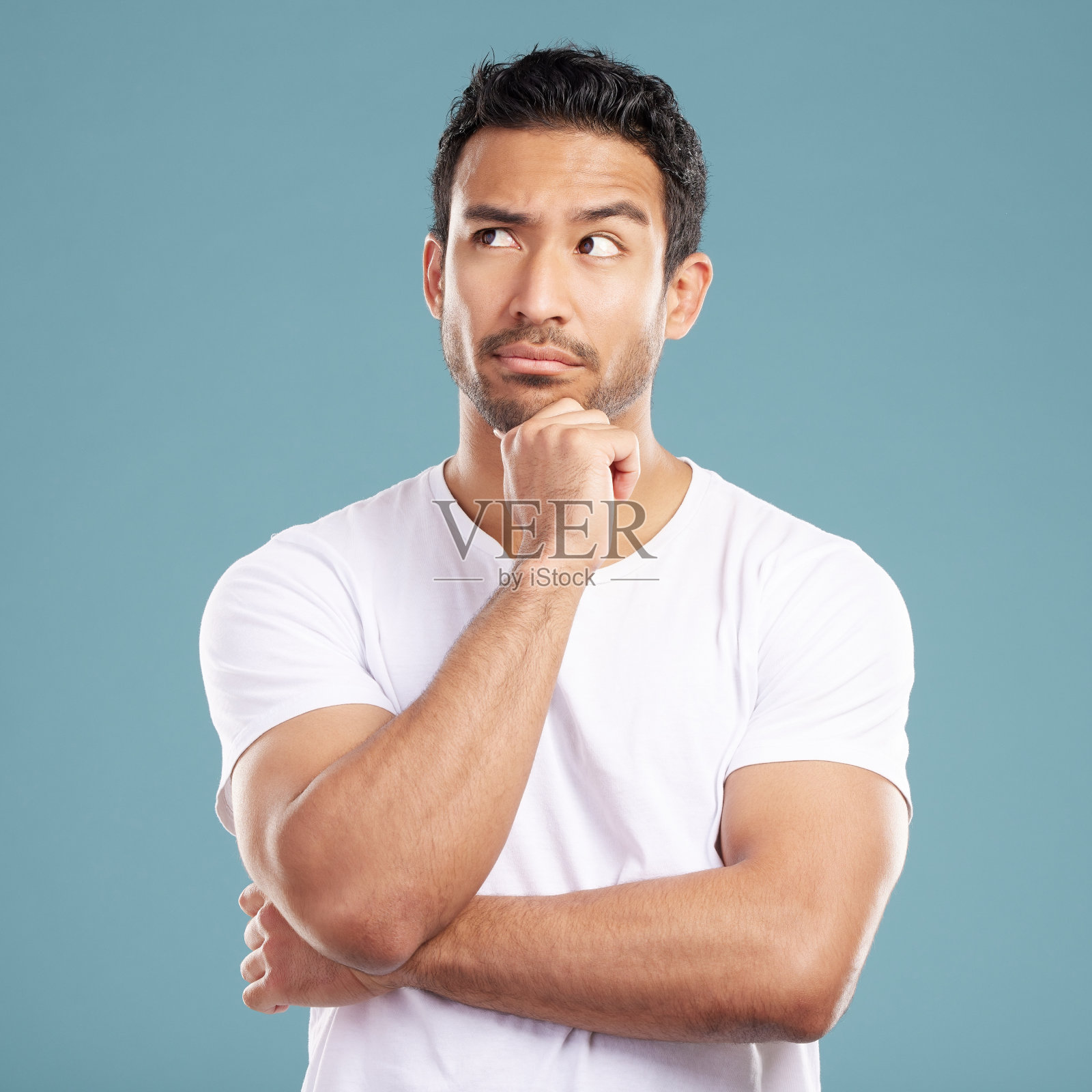 英俊的年轻混合种族男子看起来若有所思，他的手放在他的下巴和看向别处，而站在工作室孤立的蓝色背景。西班牙裔男性想到了一个好主意照片摄影图片