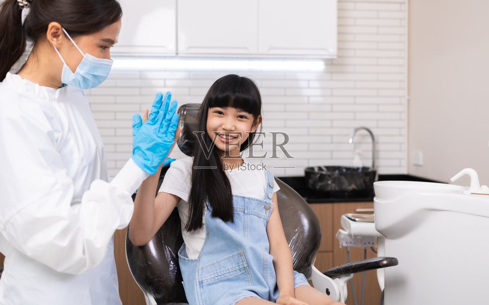 坐在椅子上的小女孩在现代设备的诊所接受牙科治疗后与儿科牙医击掌。照片摄影图片