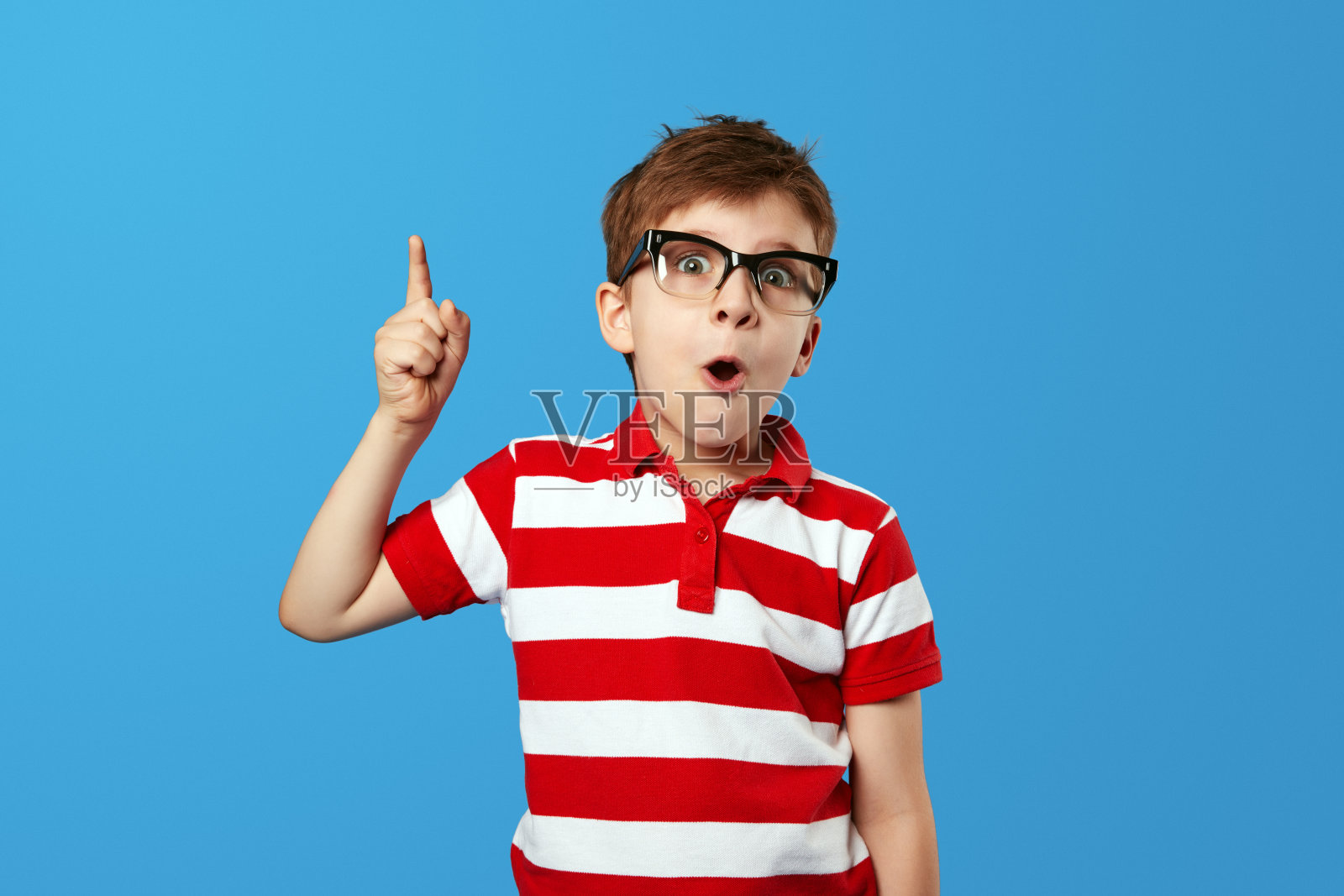 聪明的小男孩戴着眼镜，惊讶地张开嘴，指着天空，同时对蓝色有了想法照片摄影图片