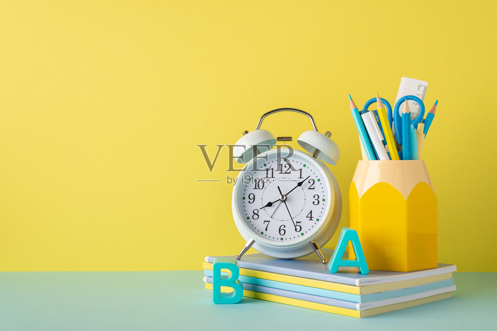 学校用品概念。黄色墙壁背景上的铅笔和塑料字母的闹钟堆叠日记的照片照片摄影图片