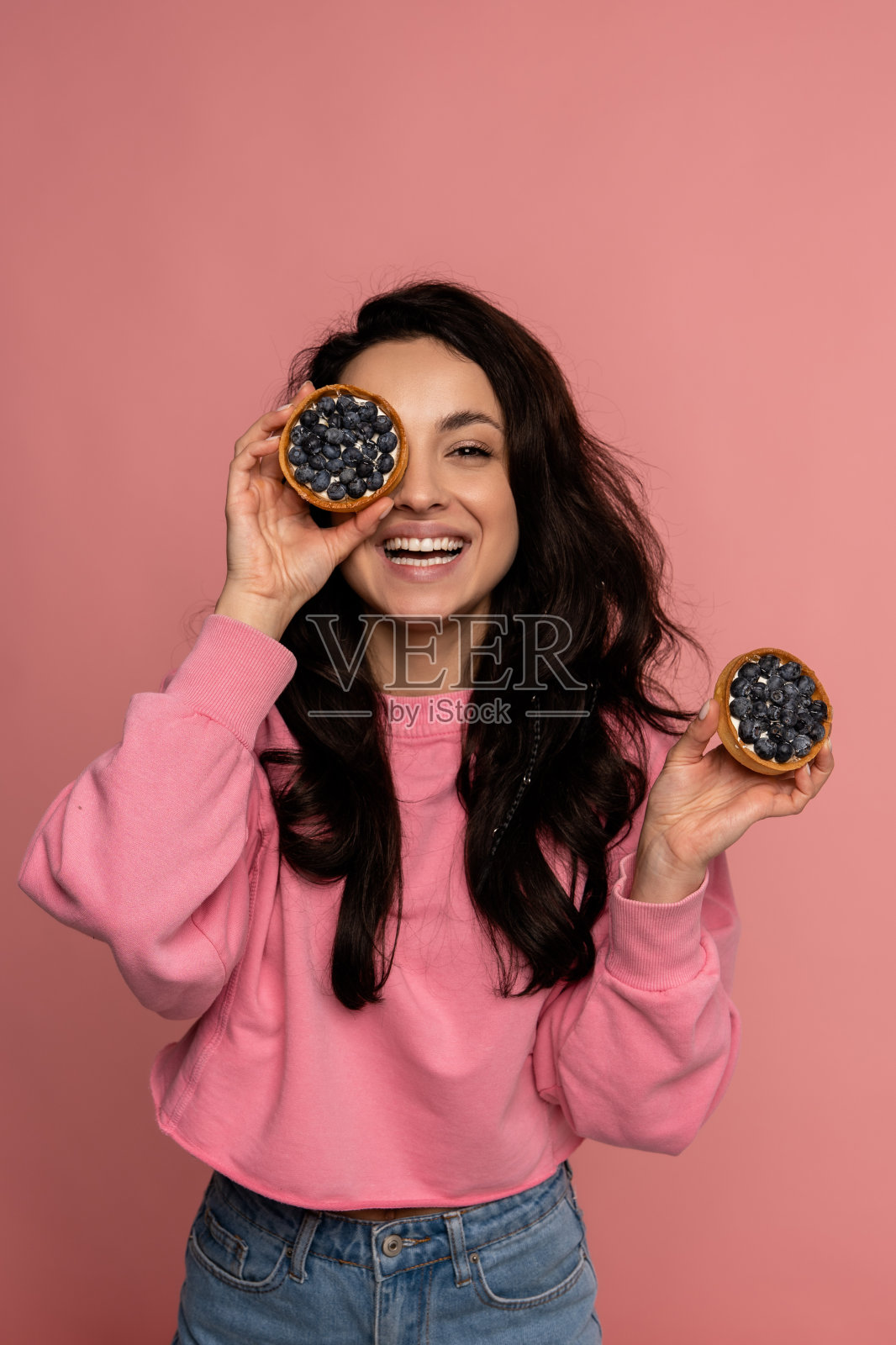 快乐的黑发年轻女性在镜头前摆姿势，粉红色的背景上有几块水果饼干。有趣又甜的食物概念照片摄影图片