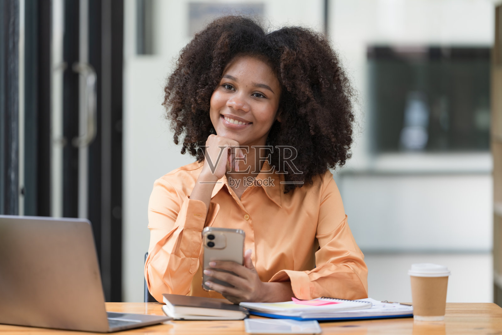 一个美丽自信的女商人坐在现代办公室里，拿着手机，拿着笔记本电脑。面带微笑的非裔美国自由职业者在家在线工作。照片摄影图片