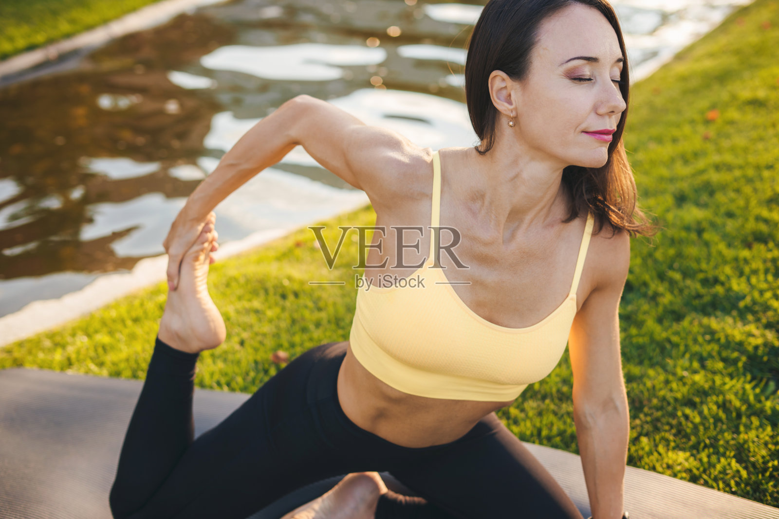 一名穿着运动服的成年女性在户外公园的健身垫上锻炼前伸展身体。健康积极的生活方式。公园户外瑜伽训练。照片摄影图片