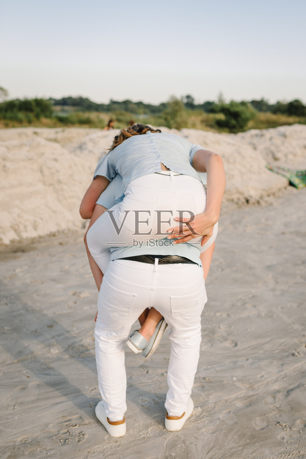 肩扛。在夏日的夕阳下，一个年轻人背着一个女人。女孩坐在沙滩上和男友拥抱在沙滩上。可爱的夫妇玩得很开心。一起度过的时光。后视图。照片摄影图片